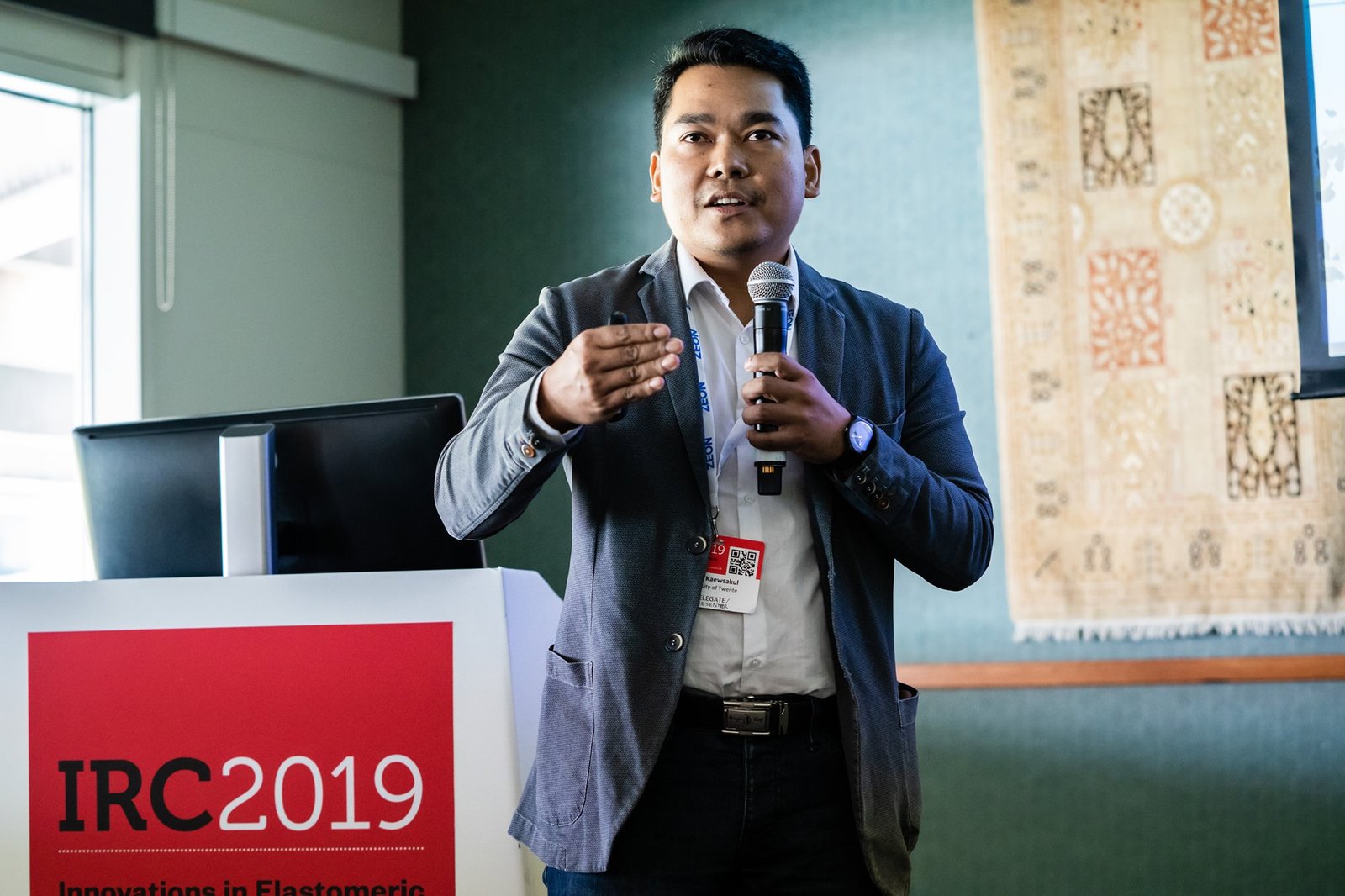 A person holding a microphone and speaking at a podium with "IRC 2019 Innovations in Elastomeric" displayed on the front. The individual is wearing a grey blazer, standing in a room with green walls and a patterned wall hanging