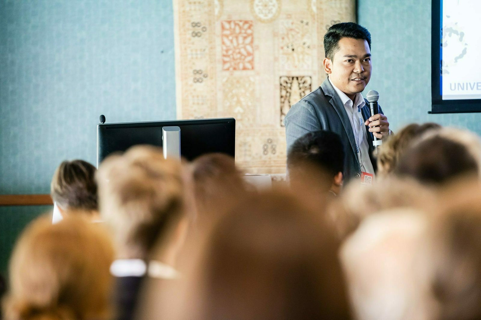 A person holding a microphone speaks to an audience in a conference room. A computer monitor is visible in the background, along with decorative wall art. The speaker wears a suit, and the room is filled with attendees. James Gifford-Mead Photography - Event Photographer London