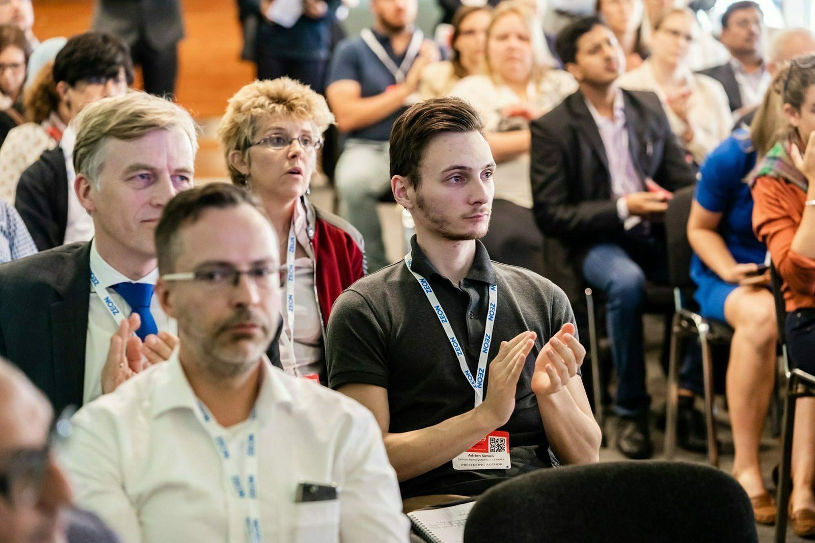 A diverse group of people sit in an audience. Several individuals are clapping, and others listen attentively. Some wear conference badges, and there is a mix of men and women dressed in business casual attire. James Gifford-Mead Photography - Event Photographer London