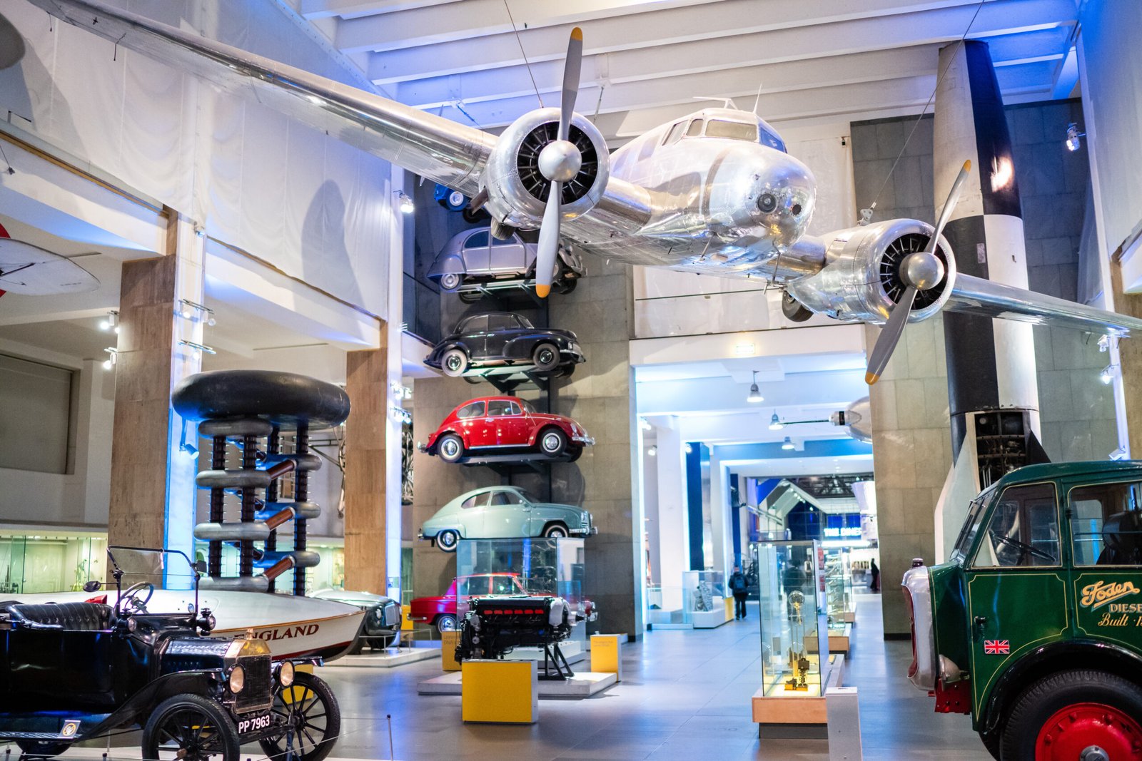 An indoor museum exhibit featuring a suspended propeller airplane. Various vintage cars and vehicles are displayed below, including a red compact car and classic vehicles. Bright lights illuminate the spacious hall