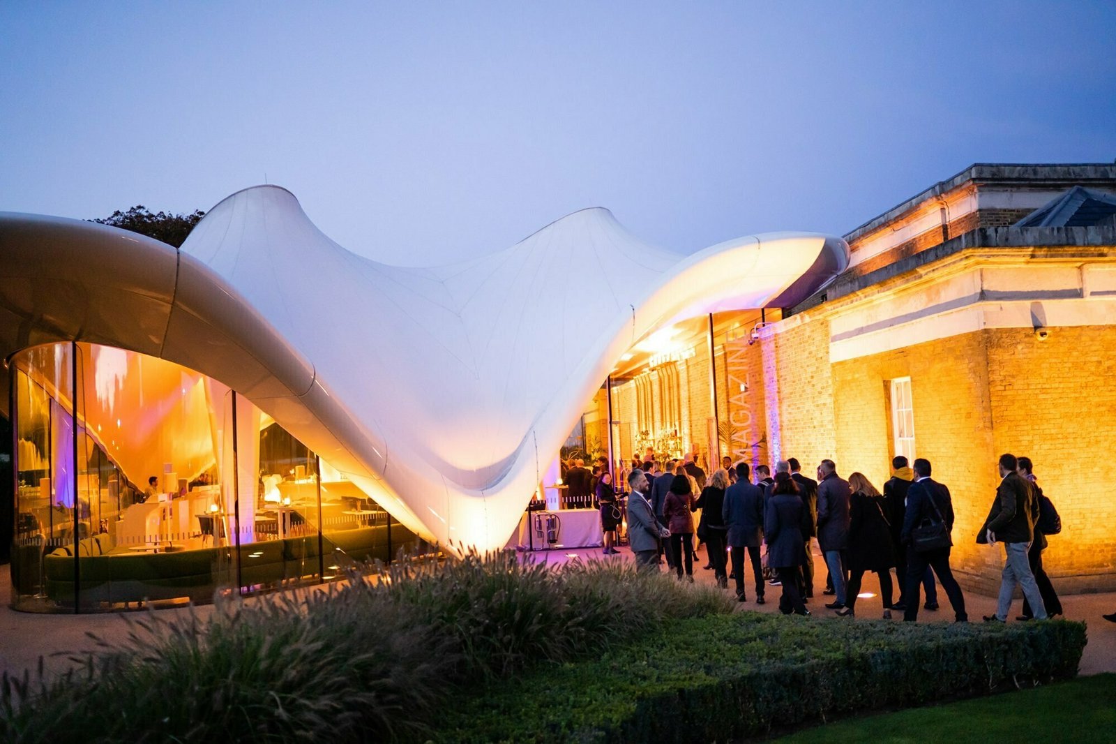 A modern architectural building with a flowing white canopy is illuminated in various colors. People are gathered outside, socializing near the entrance in the evening. James Gifford-Mead Photography - Event Photographer London