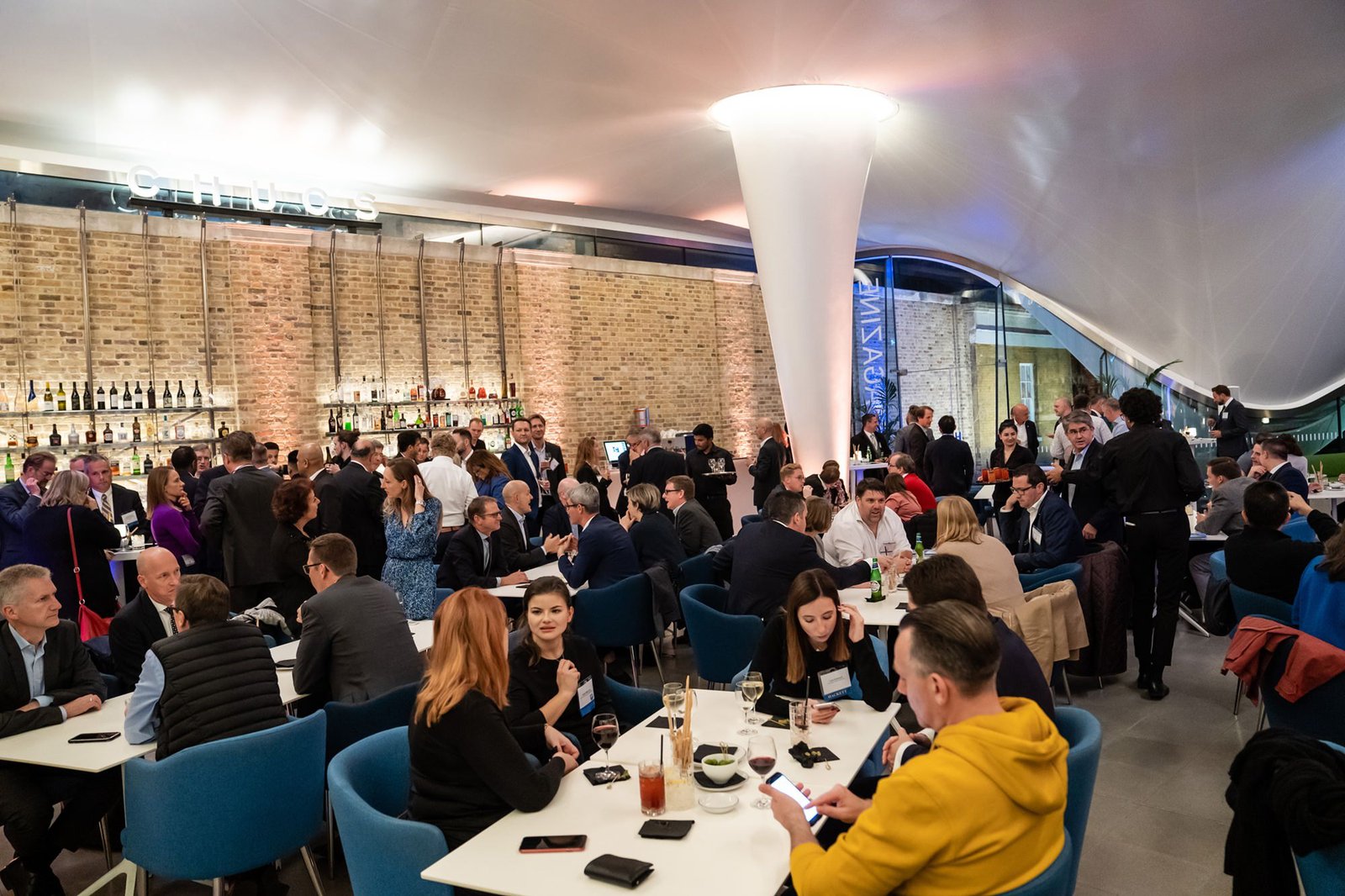A lively, modern restaurant with numerous people seated at tables, engaged in conversation. The space features a brick wall with a backlit bar and shelves of bottles. A mix of casually and formally dressed patrons adds to the energetic ambiance. James Gifford-Mead Photography - Event Photographer London