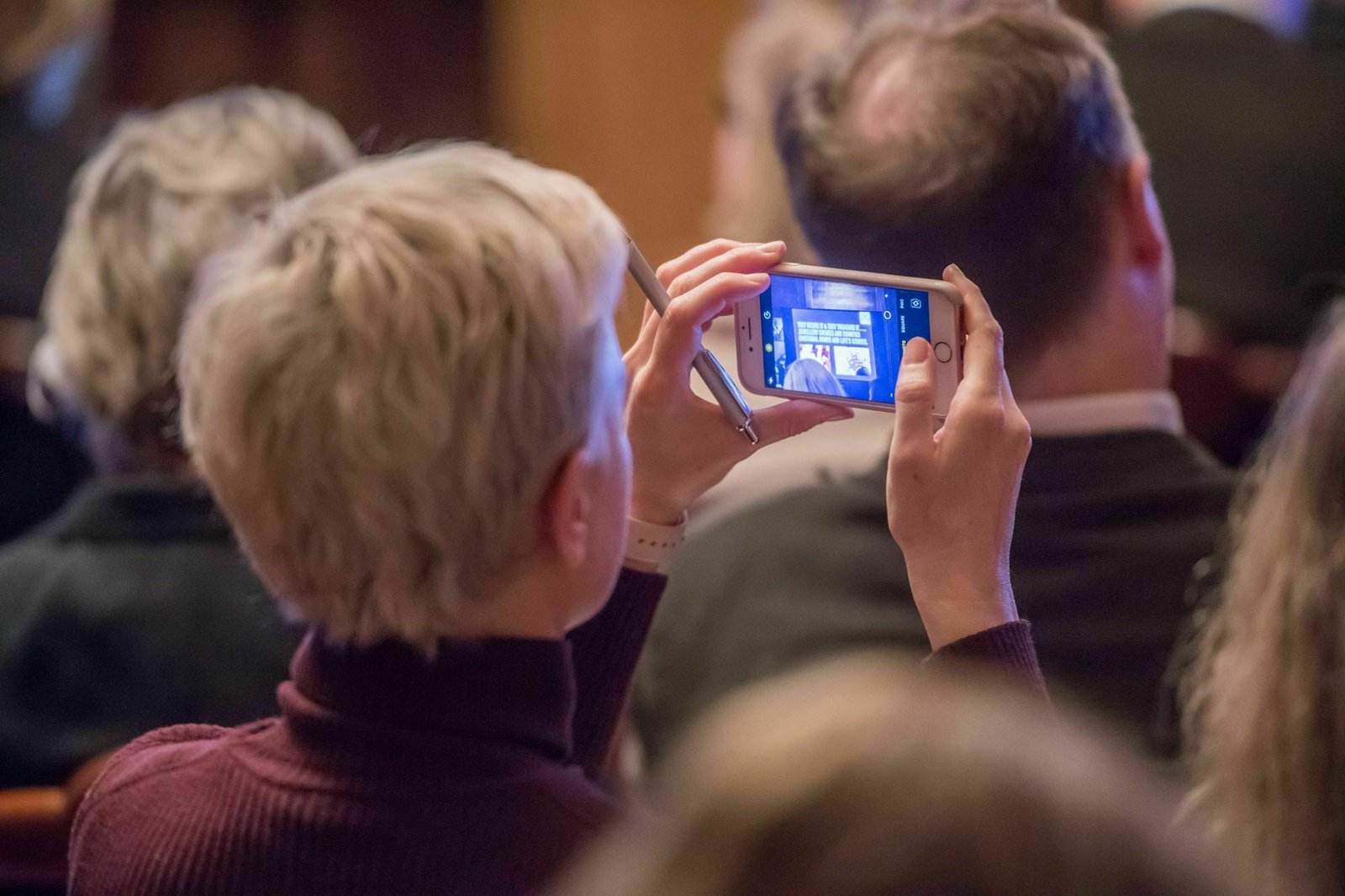 A person with short blonde hair is sitting among a group of people. They are holding a smartphone horizontally, capturing a photo or video of something in front of them. The scene appears to be in a formal or crowded indoor setting. James Gifford-Mead Photography - Event Photographer London
