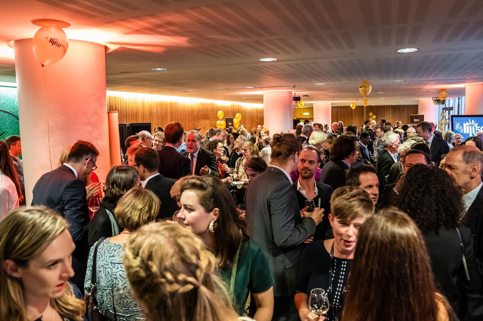 A crowded indoor event with people socializing and holding drinks. Attendees are dressed in formal and business attire. The room is warmly lit, with white pillars and golden balloons, creating a festive atmosphere