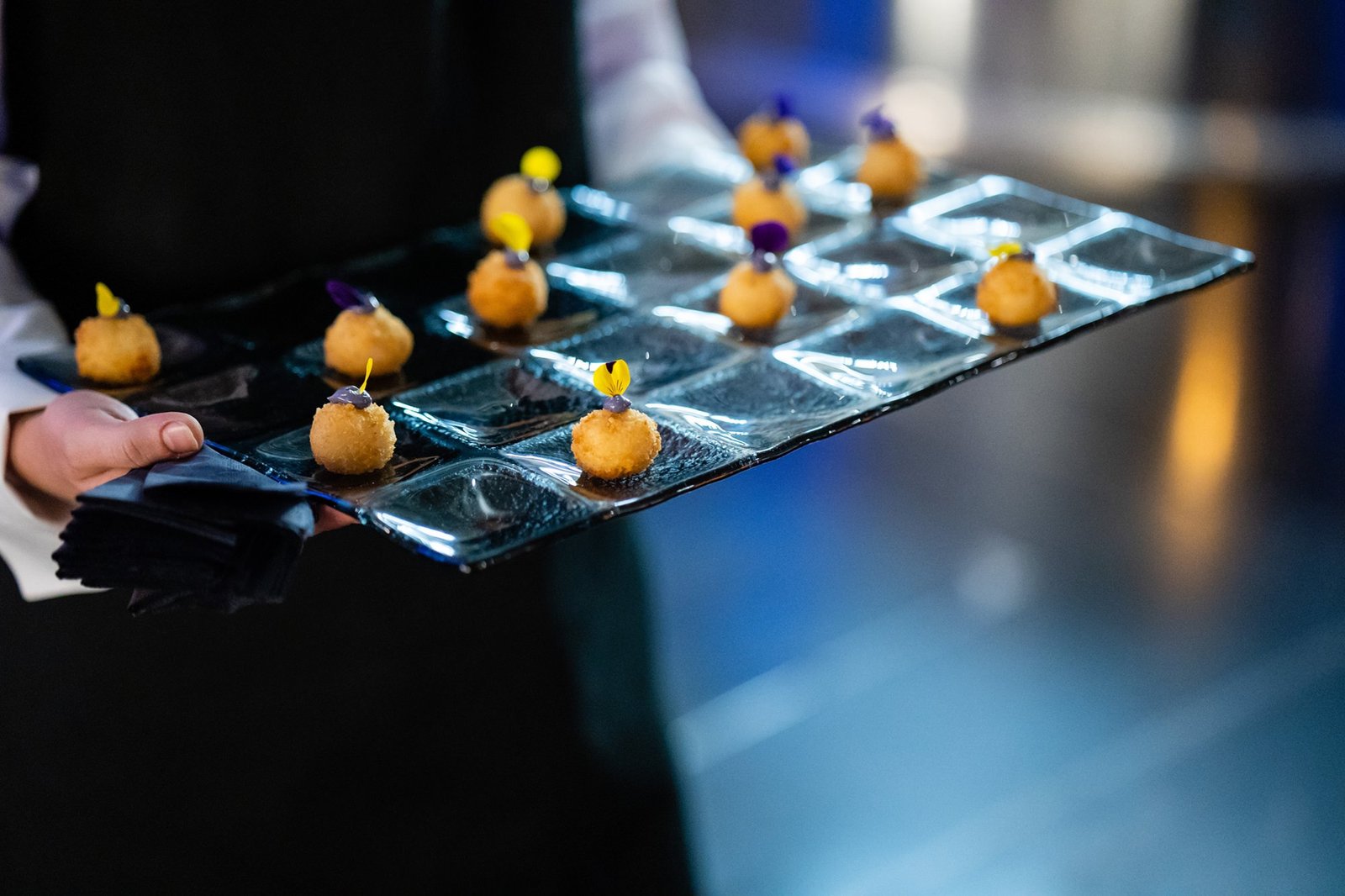 A person holds a black tray with multiple bite-sized appetizers, each garnished with a small yellow or purple flower, arranged in individual sections. The background is softly blurred. James Gifford-Mead Photography - Event Photographer London