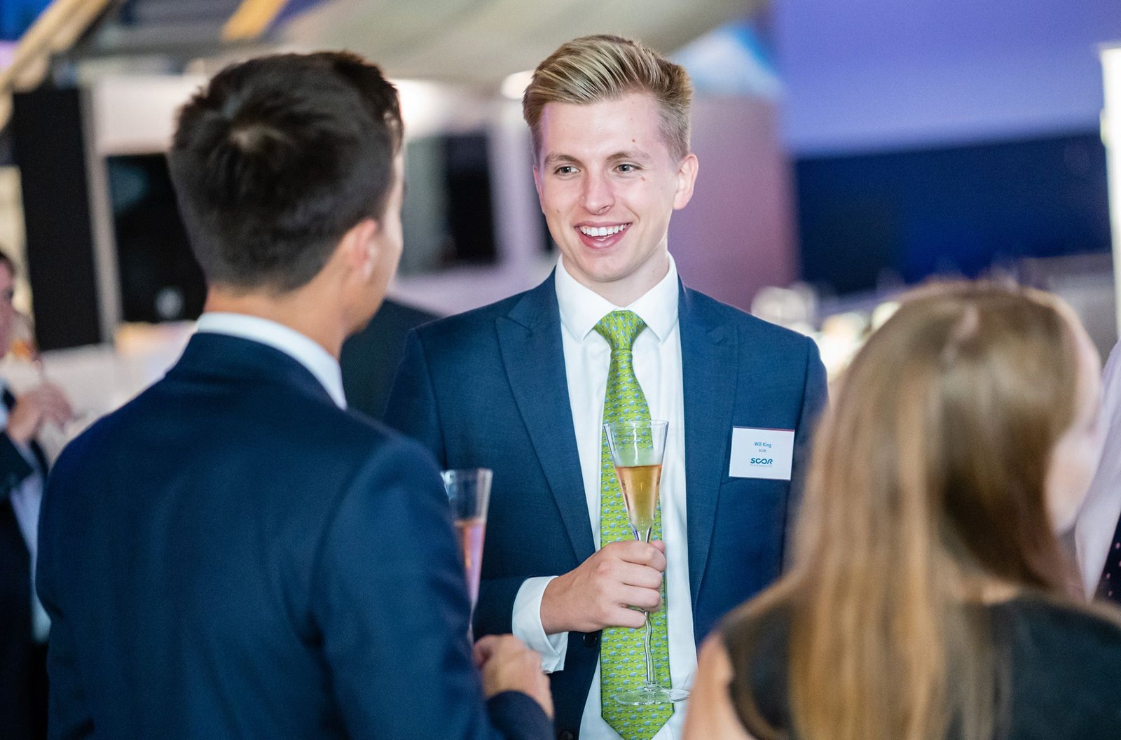 A young man in a suit with a green tie is holding a champagne glass and smiling while conversing with others at an event. Another person in a suit faces him, and a woman with long hair is partially visible. James Gifford-Mead Photography - Event Photographer London