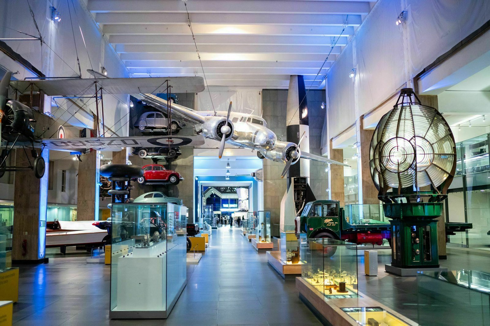 A spacious museum gallery displaying vintage aircraft and vehicles. Glass cases exhibit small models and artifacts. Modern lighting highlights a large plane suspended from the ceiling, with various historical items showcased around the floor. James Gifford-Mead Photography - Event Photographer London