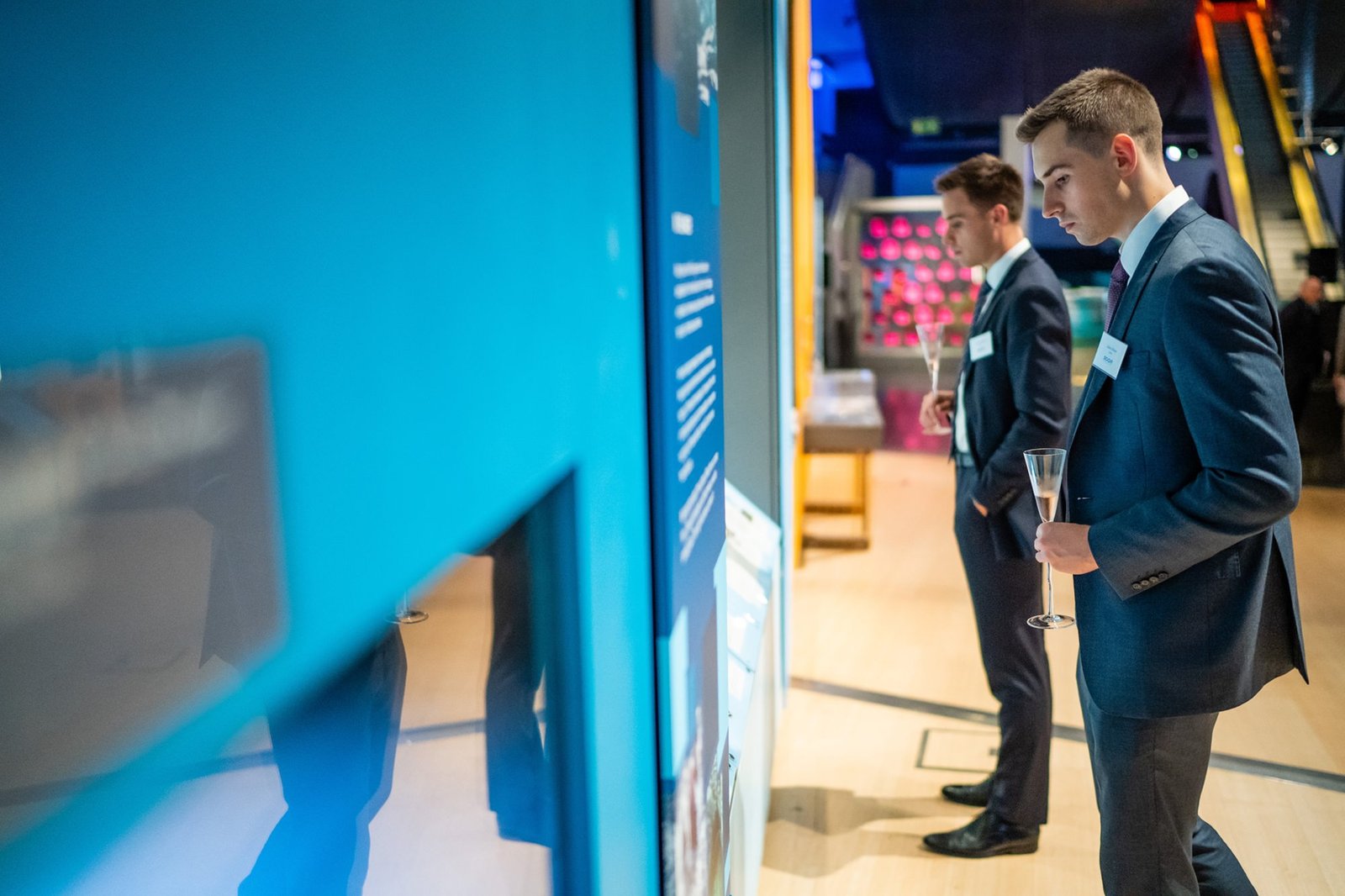 Two men in suits stand in an exhibition space, looking at a display. One holds a champagne flute. The background features colorful lighting and various exhibits. The setting appears formal and modern. James Gifford-Mead Photography - Event Photographer London