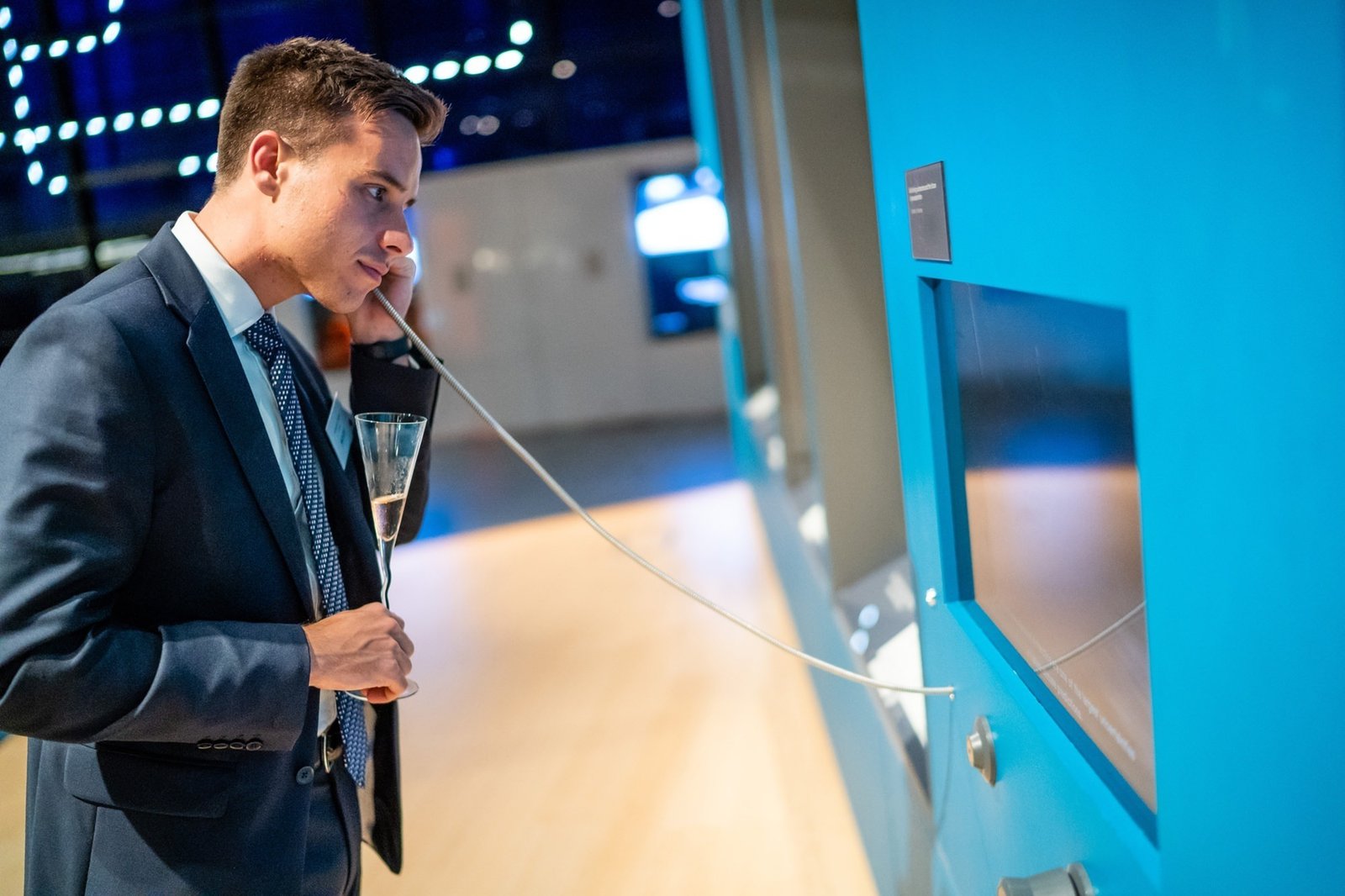 A man in a blue suit and tie examines a display at an exhibition. He holds a glass of champagne and leans in, looking intently at the object inside. The background is softly lit and features modern decor elements. James Gifford-Mead Photography - Event Photographer London