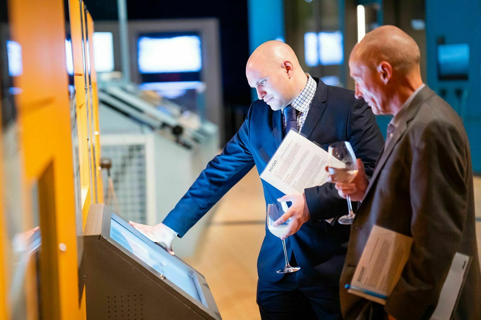 Two men in business attire interact with a touchscreen kiosk. One points at the screen while holding papers and a glass of wine. The other observes, holding a book and a wine glass. They are in a brightly lit, modern indoor setting. James Gifford-Mead Photography - Event Photographer London