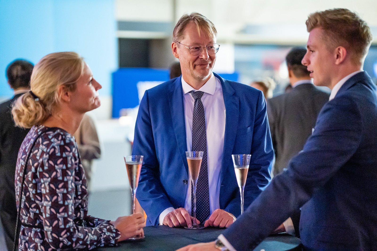A woman and two men in formal attire stand around a high table with champagne glasses, engaged in conversation. The setting appears to be a social or corporate event, with other people in the blurred background. James Gifford-Mead Photography - Event Photographer London