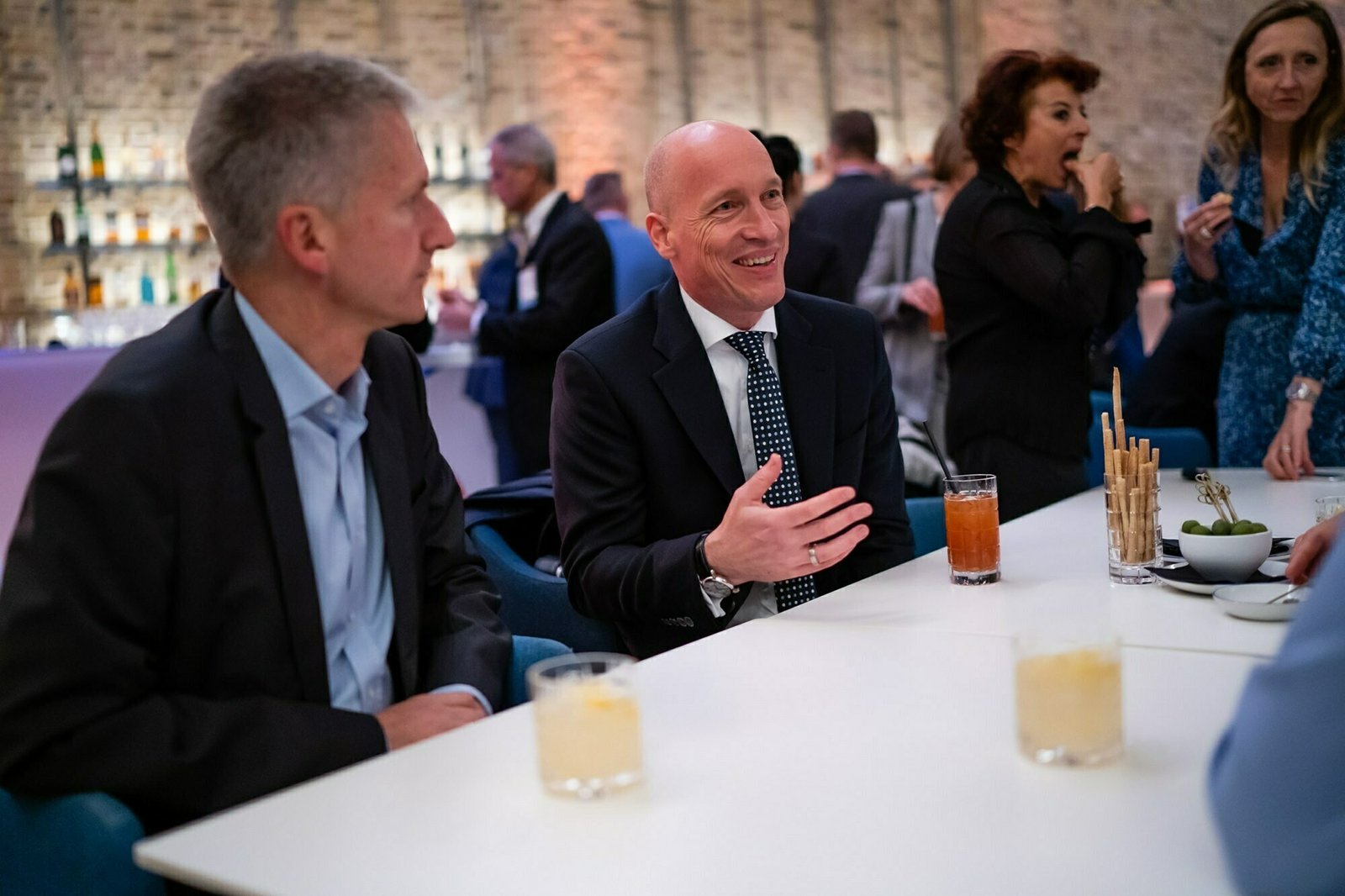 Two men in suits sit at a table with drinks, engaged in conversation. Behind them, people stand and chat in a warmly lit room. The setting appears to be a social or networking event. James Gifford-Mead Photography - Event Photographer London