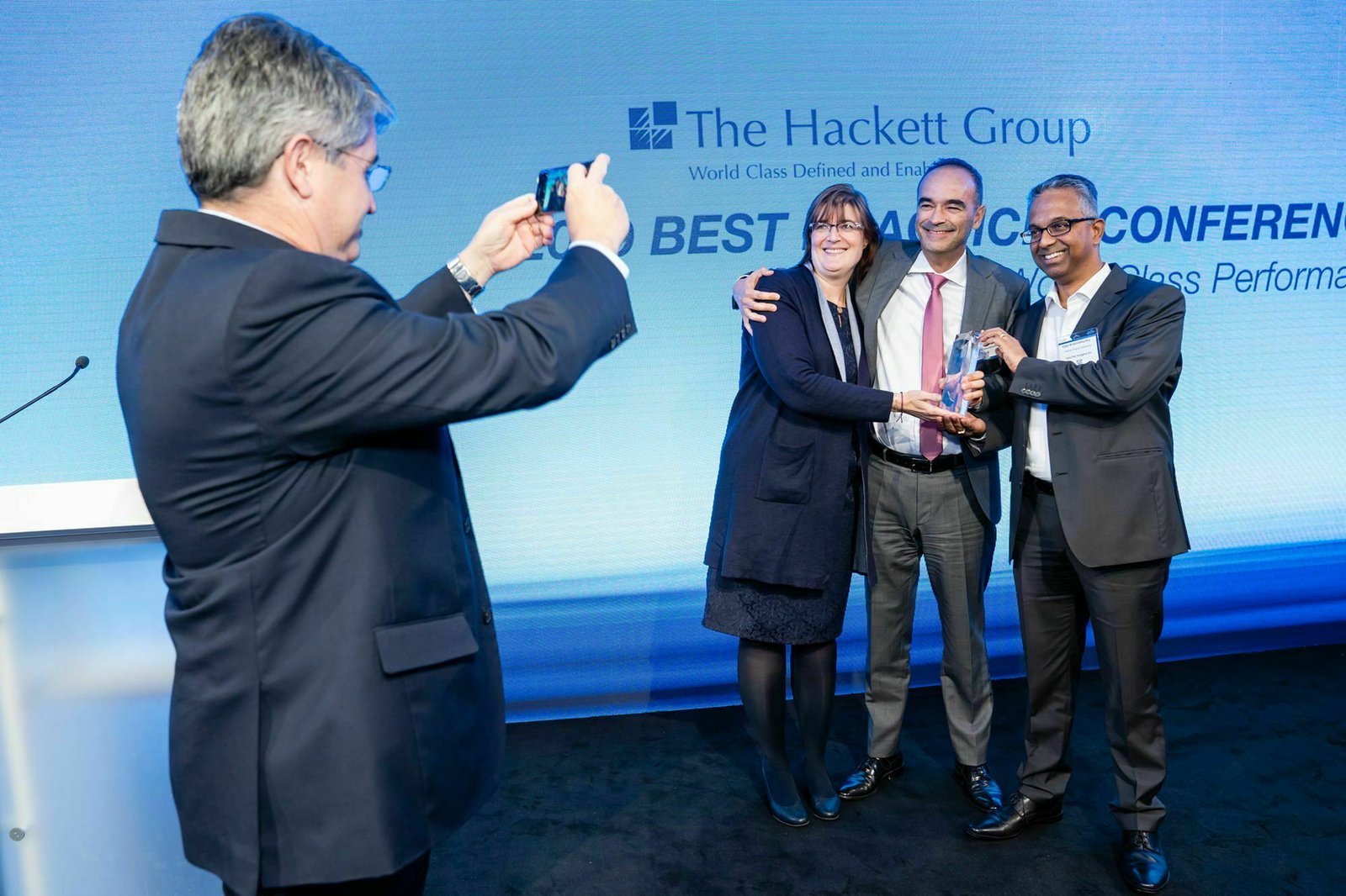 A man takes a photo of two men and a woman smiling and holding an award on stage at The Hackett Group event. The backdrop reads "Best Practices Conference. James Gifford-Mead Photography - Event Photographer London
