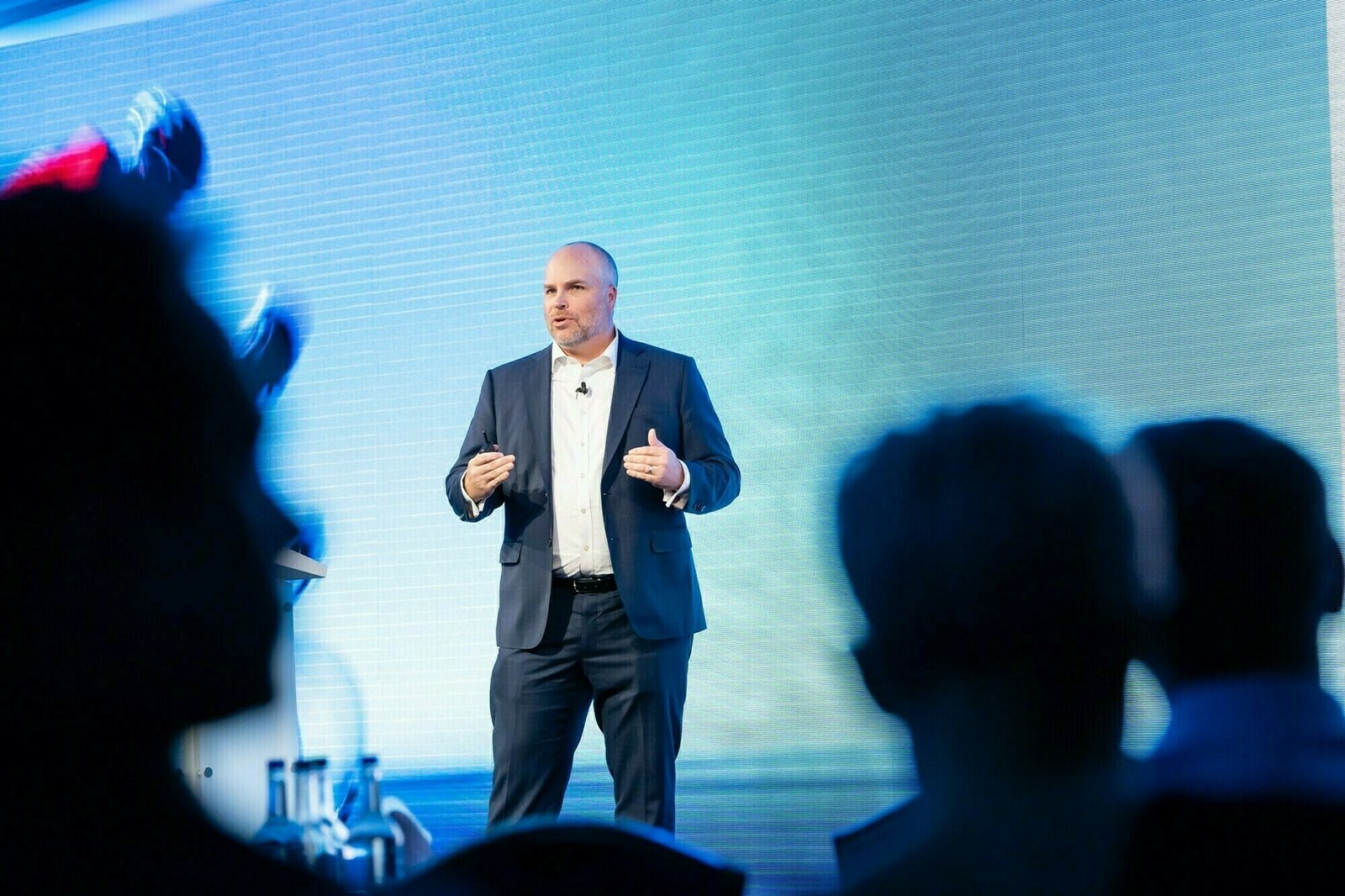 A man in a suit and open-collared shirt speaks on stage in front of a large screen displaying a blue image. Audience members are silhouetted in the foreground. James Gifford-Mead Photography - Event Photographer London