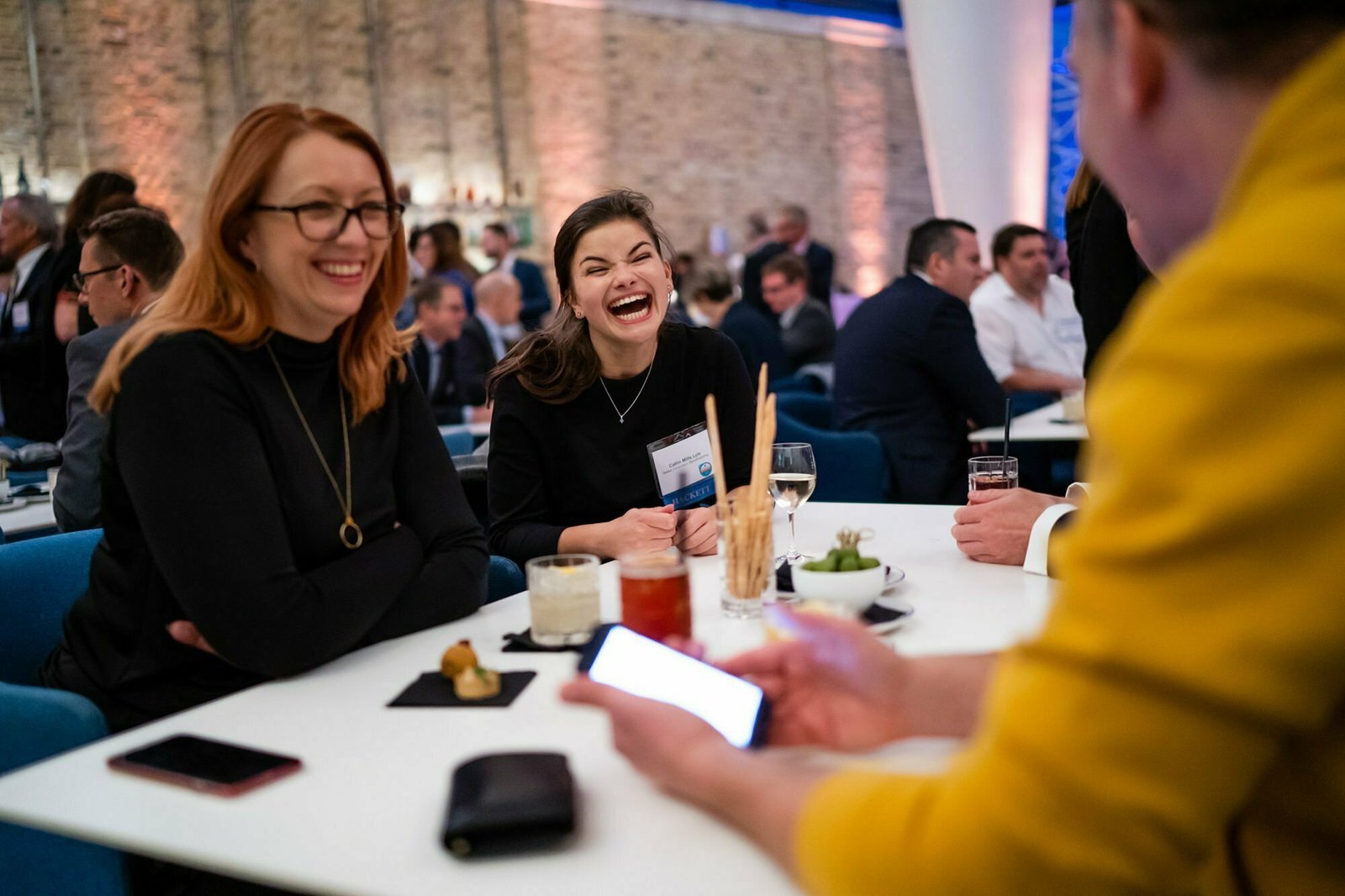 Three people are sitting at a table, enjoying a lively conversation and laughing. The setting appears to be a social or networking event, with other people visible in the background. They have drinks and snacks on the table. James Gifford-Mead Photography - Event Photographer London