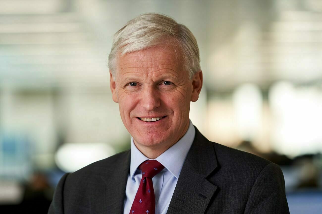 A middle-aged man with gray hair smiles, wearing a dark suit, white shirt, and red patterned tie. The background is a softly blurred indoor setting, suggesting an office environment. James Gifford-Mead Photography - Event Photographer London