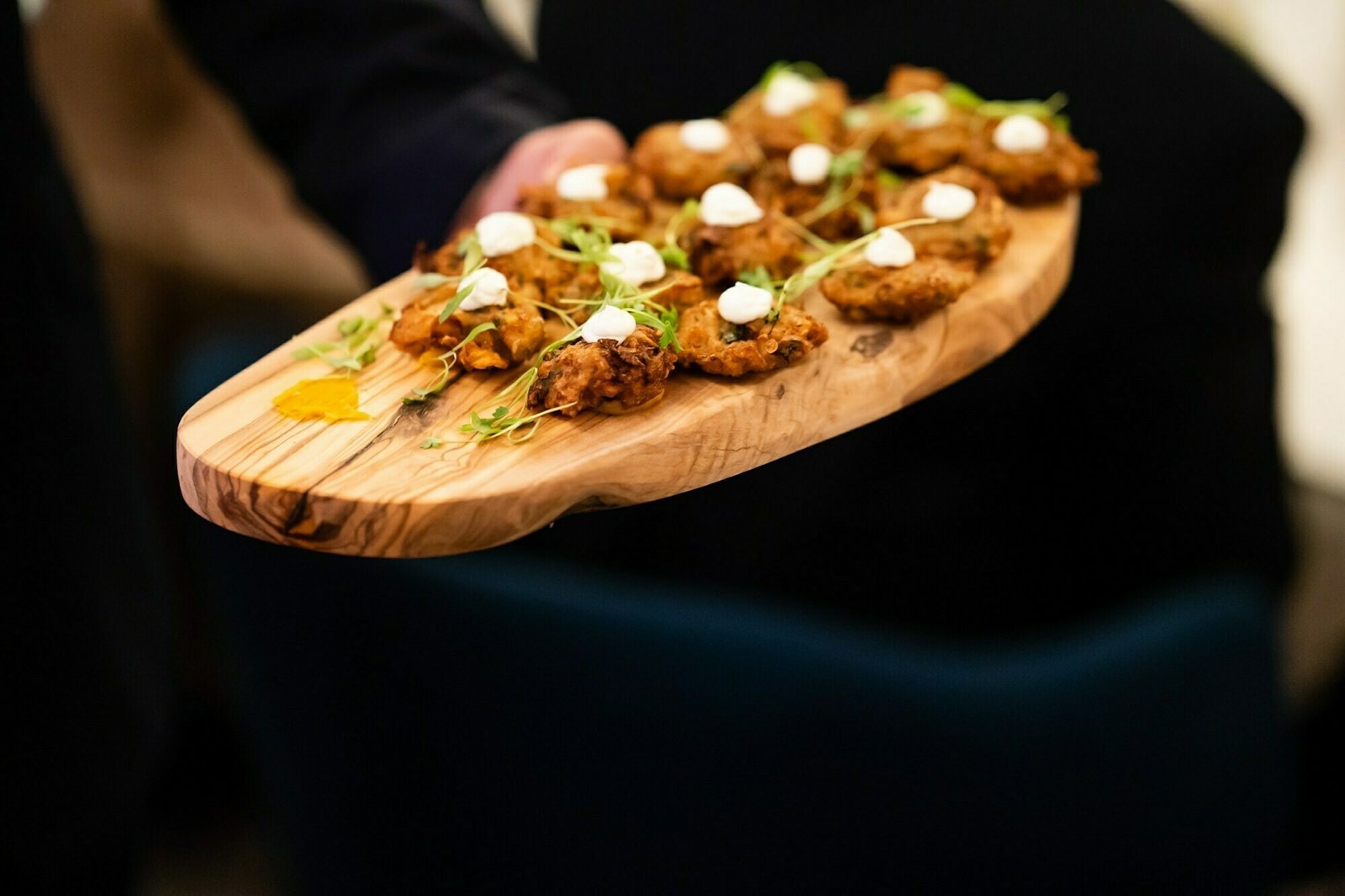 A hand holds a wooden board with several small, fried appetizers topped with white sauce and garnished with green herbs, ready to be served. The background is blurred. James Gifford-Mead Photography - Event Photographer London
