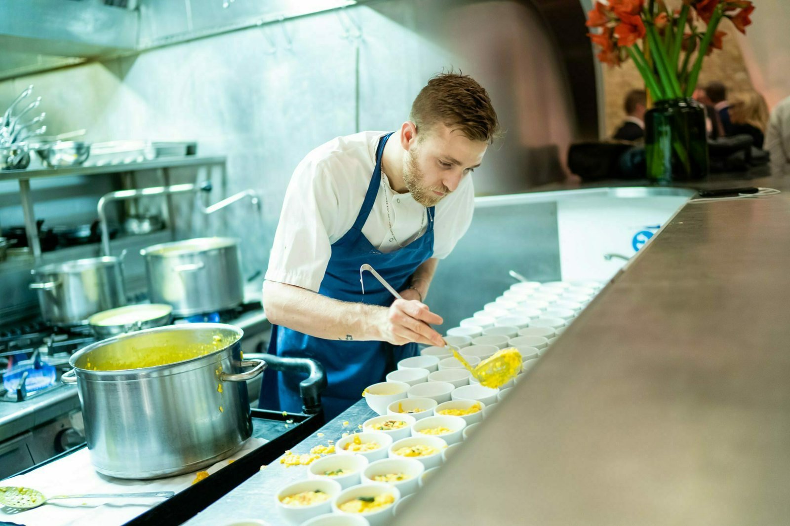A chef in a blue apron ladles soup into rows of small white bowls in a professional kitchen. A large pot of soup sits on the stove, and colorful flowers are in a vase nearby. James Gifford-Mead Photography - Event Photographer London