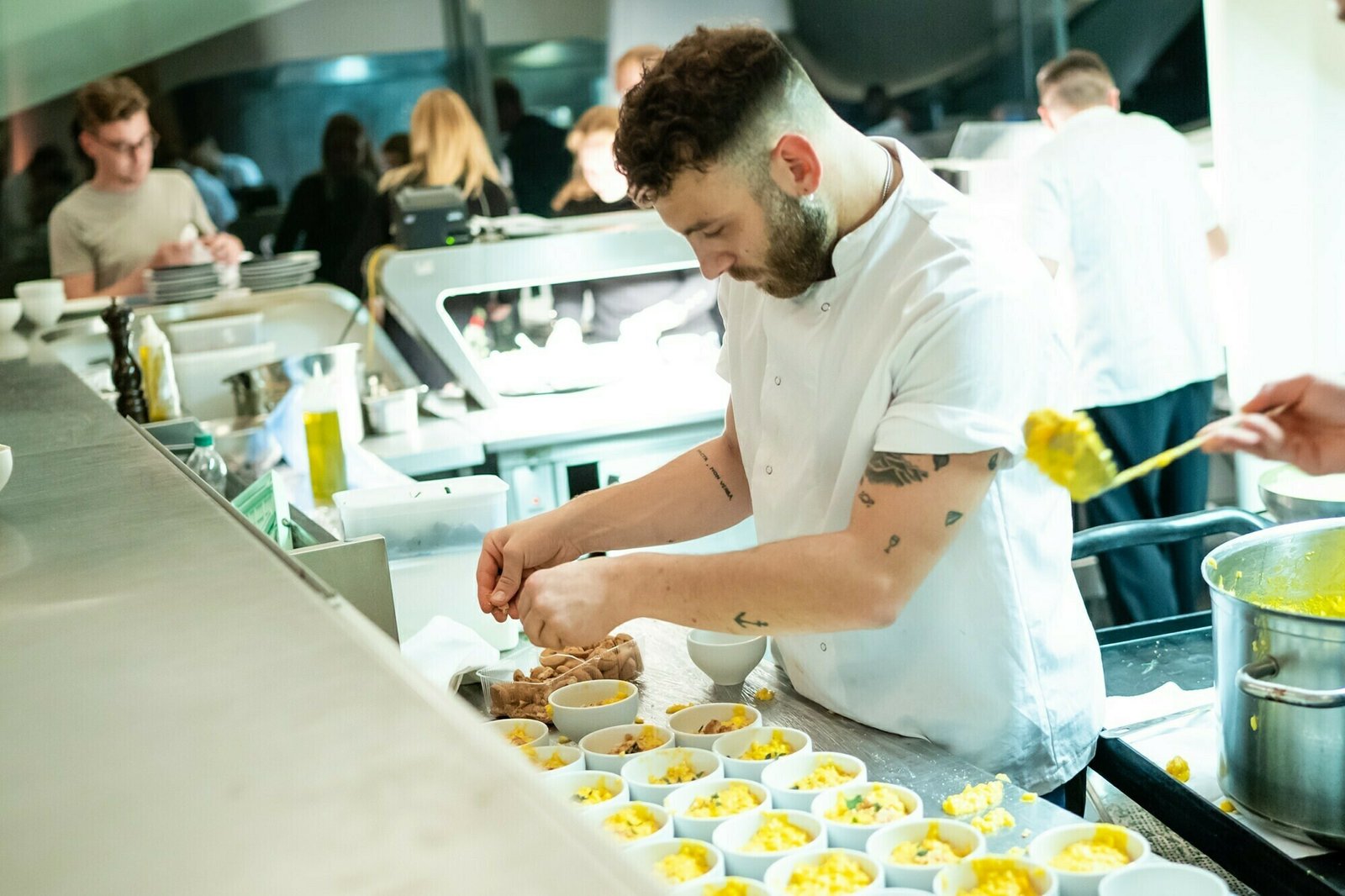 A chef with tattoos prepares small bowls of yellow rice in a busy restaurant kitchen. Other staff members work in the background, focusing on various tasks under bright lighting. James Gifford-Mead Photography - Event Photographer London