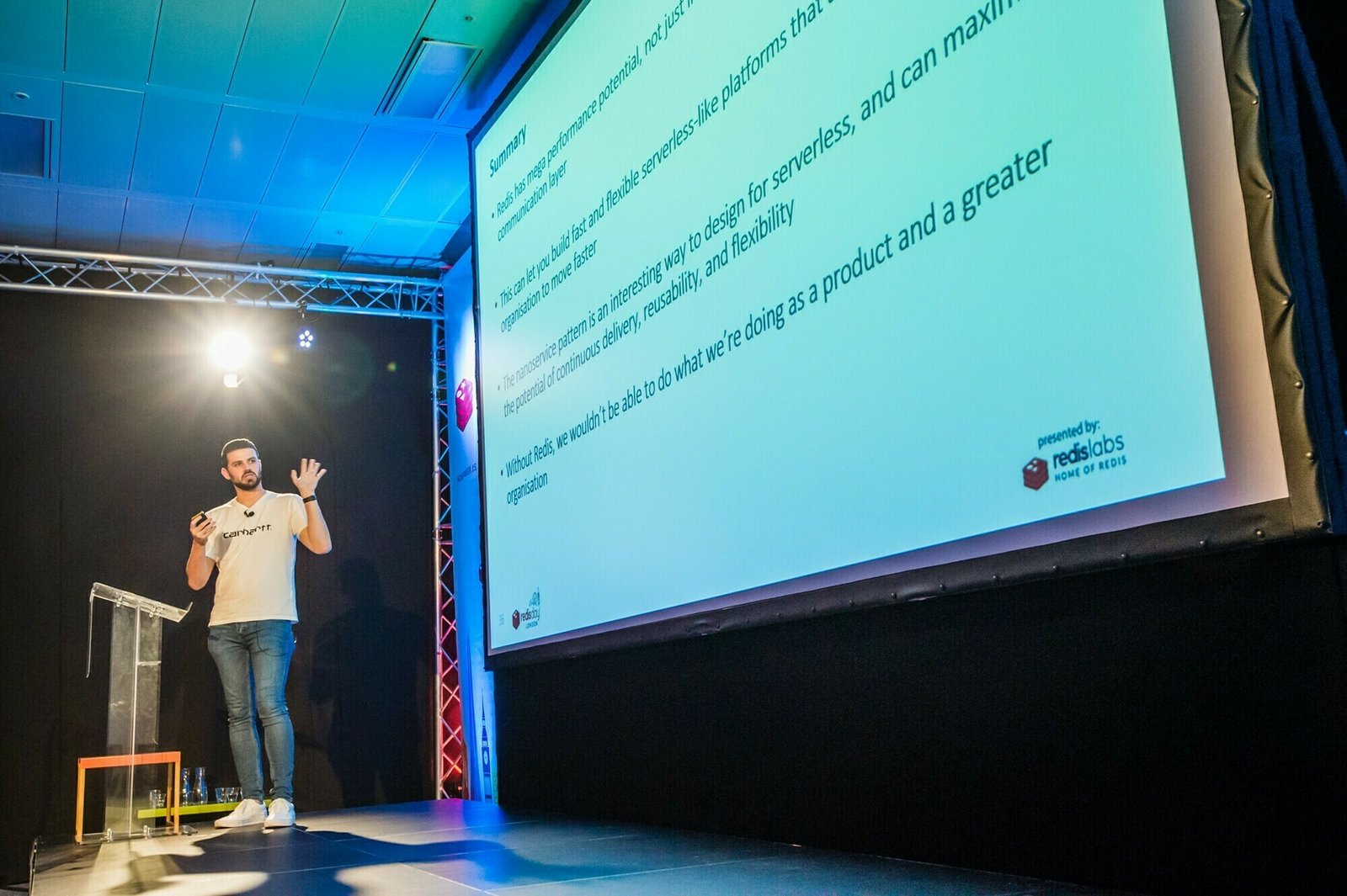 A man stands on stage during a presentation, gesturing with one hand. A large screen to his right displays a slide with text. The stage is lit with blue and purple lighting, and the audience seating is not visible. James Gifford-Mead Photography - Event Photographer London