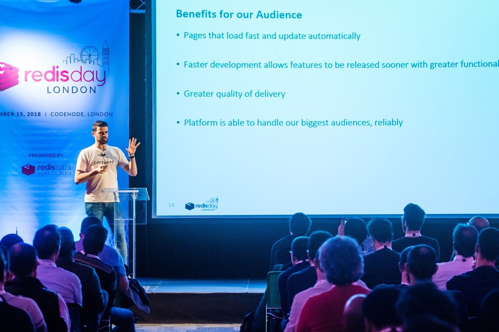 A speaker presents at a conference in London, discussing audience benefits with a slide listing points such as fast updates, development speed, quality delivery, and reliable platform handling. The audience listens attentively in a dimly lit room. James Gifford-Mead Photography - Event Photographer London