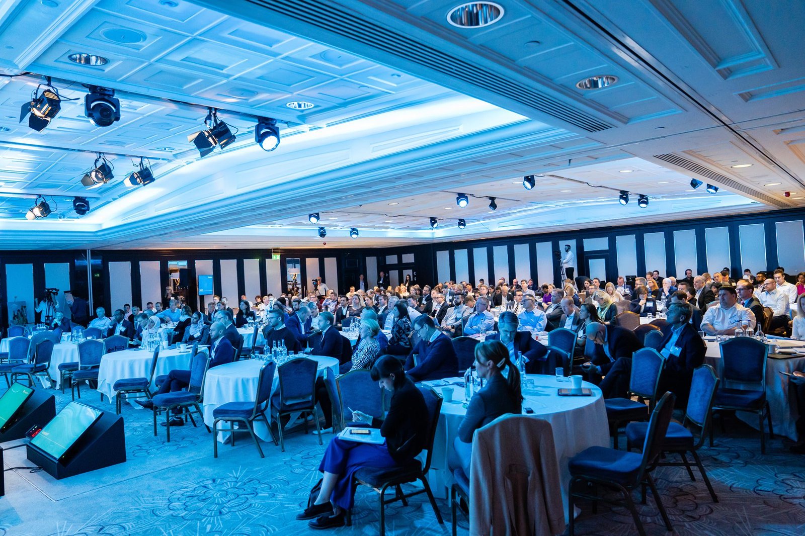 A large audience seated in a conference hall with blue lighting listens attentively to a presentation. Circular tables and rows of chairs fill the room, and stage lights hang from the ceiling. The atmosphere is professional and focused. James Gifford-Mead Photography - Event Photographer London