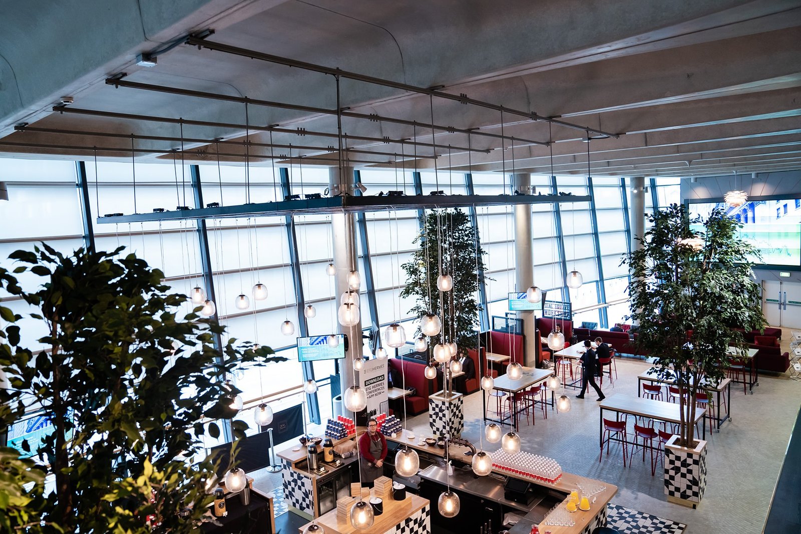 A modern cafe with hanging lights and tall plants. The space features red high tables, chairs, and a counter with patterned tiling. Large windows allow natural light inside. Two people walk in the background. James Gifford-Mead Photography - Event Photographer London