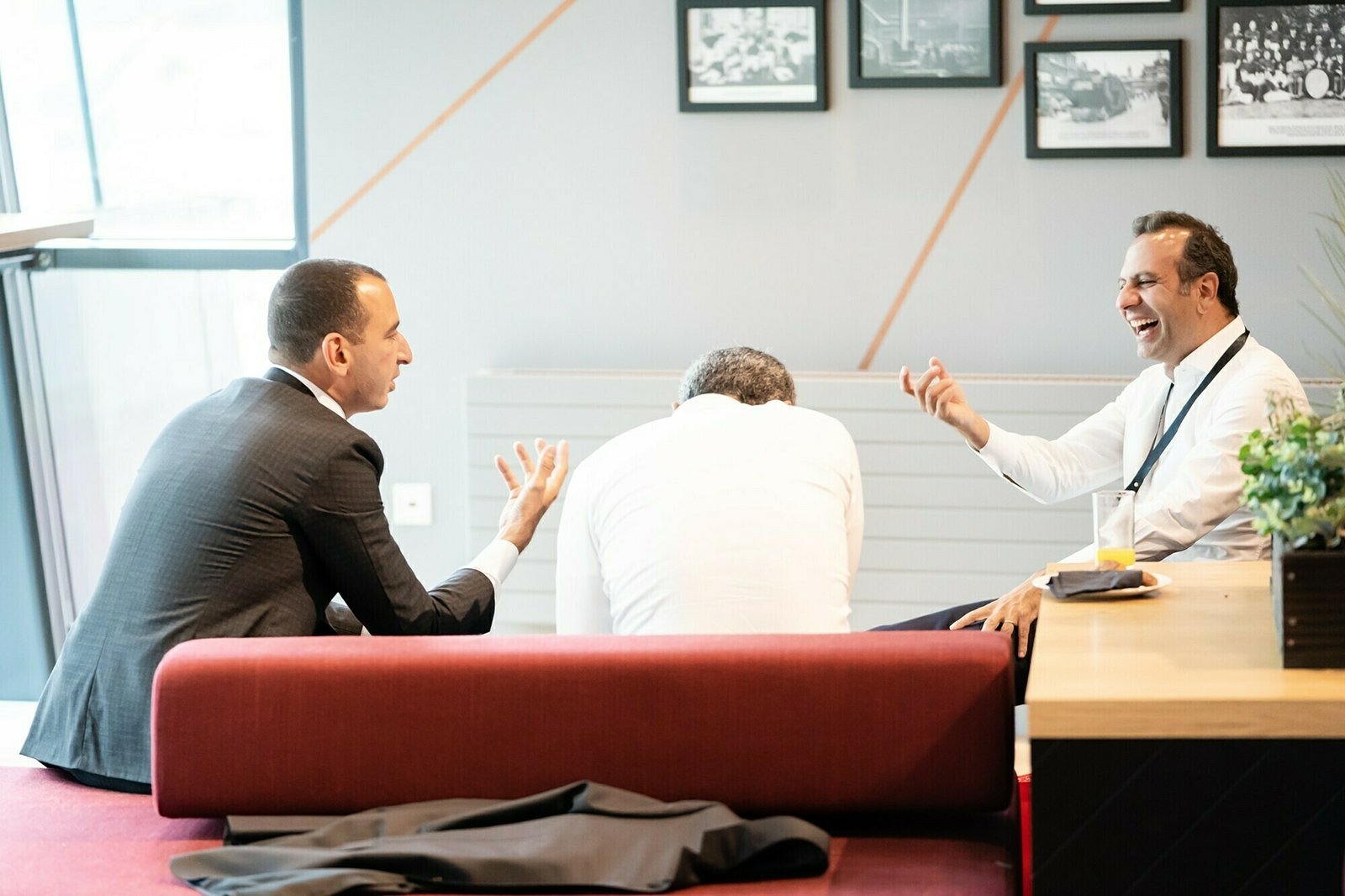 Three men in formal attire sit on a red bench engaging in a lively discussion. Two are animatedly gesturing, while the third leans forward. Framed photos adorn the wall behind them. James Gifford-Mead Photography - Event Photographer London