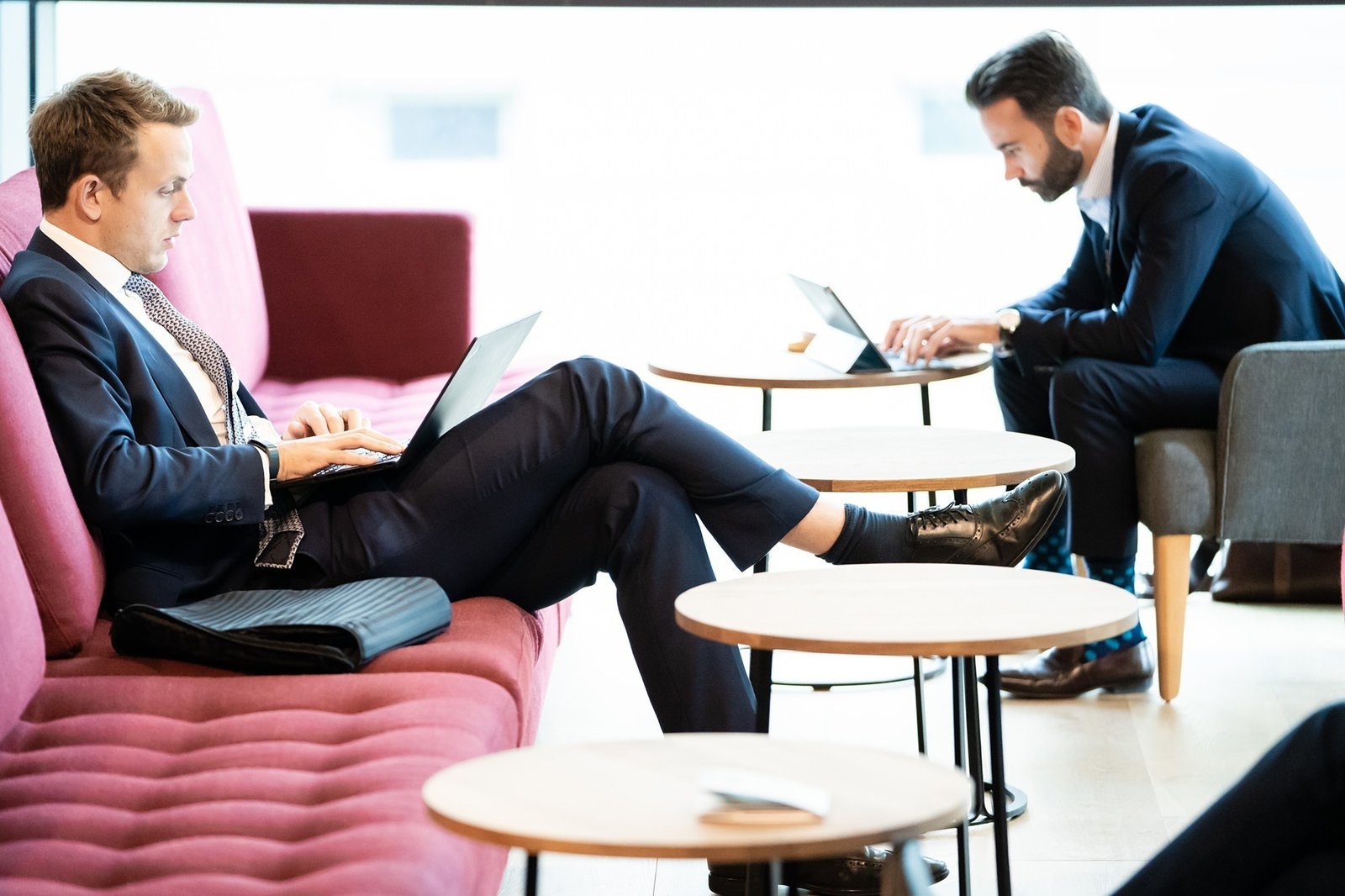 Two men in suits are working on laptops. One is sitting on a maroon sofa with crossed legs, while the other is at a small table in a gray chair. The setting is a modern lounge with wooden tables and a bright window. James Gifford-Mead Photography - Event Photographer London
