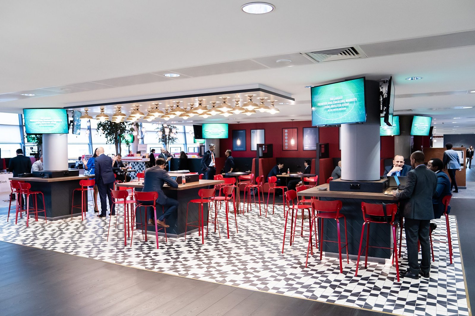 A modern lounge with red chairs and black bar counters, tiled floor, and multiple screens. People are sitting and standing, engaging in conversations. The setting is well-lit with a casual and social atmosphere. James Gifford-Mead Photography - Event Photographer London