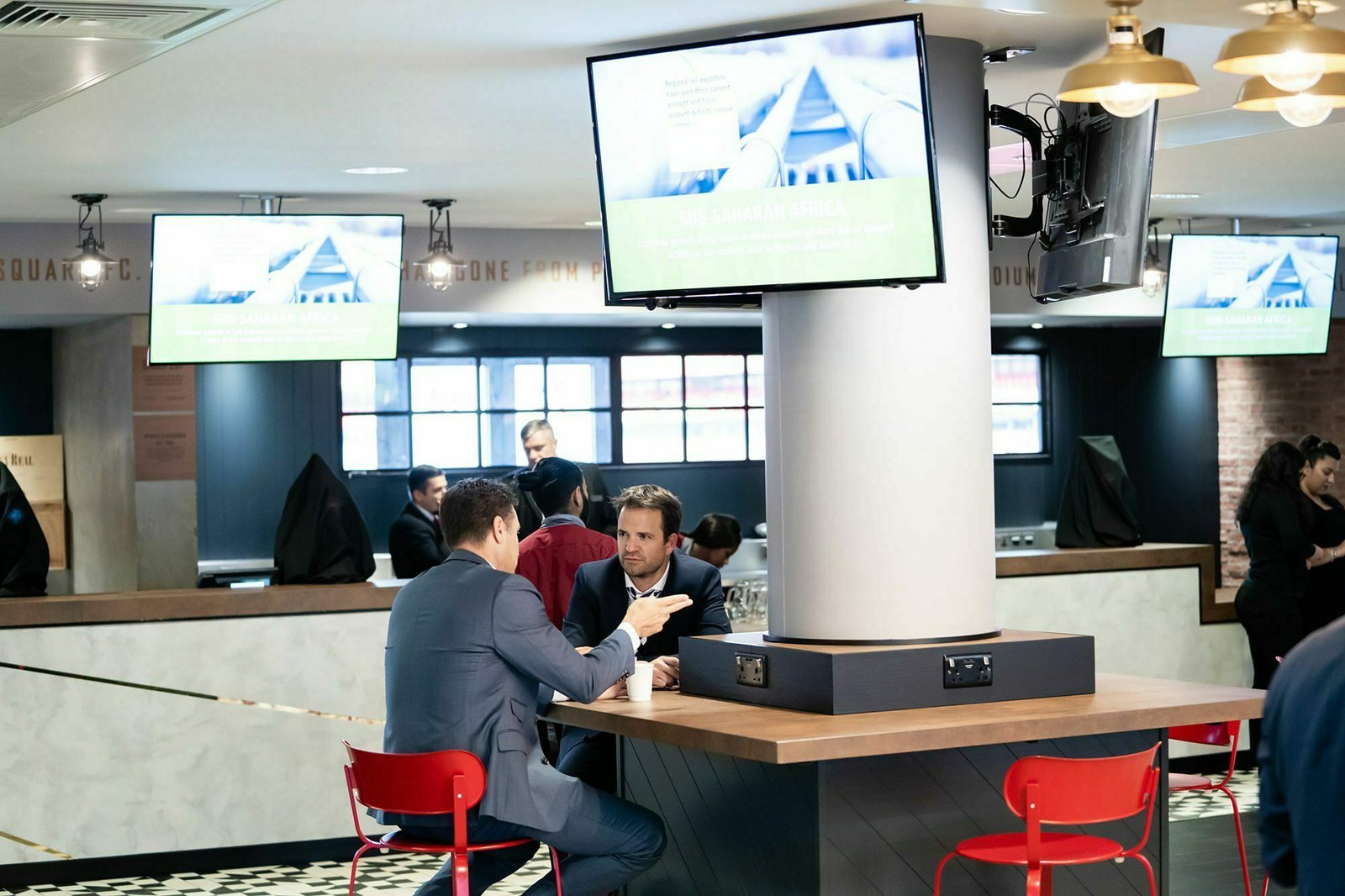 Two people sitting at a high-top table talking in a modern cafe or restaurant. A TV screen displays information above them. The decor includes red chairs, a white pillar, and soft overhead lighting. Other patrons and staff are seen in the background. James Gifford-Mead Photography - Event Photographer London