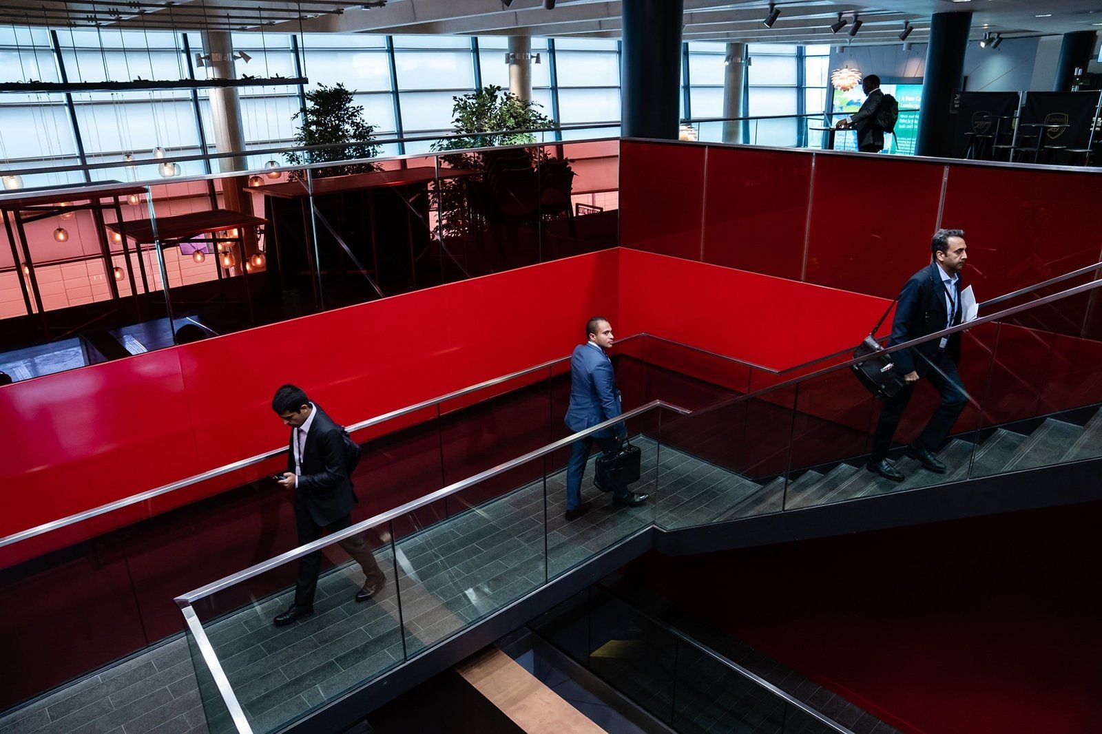 Three men in business attire walk through a modern, brightly lit building with red walls. Two use a staircase, while one walks below. They carry bags, and potted plants are visible near large windows in the background. James Gifford-Mead Photography - Event Photographer London