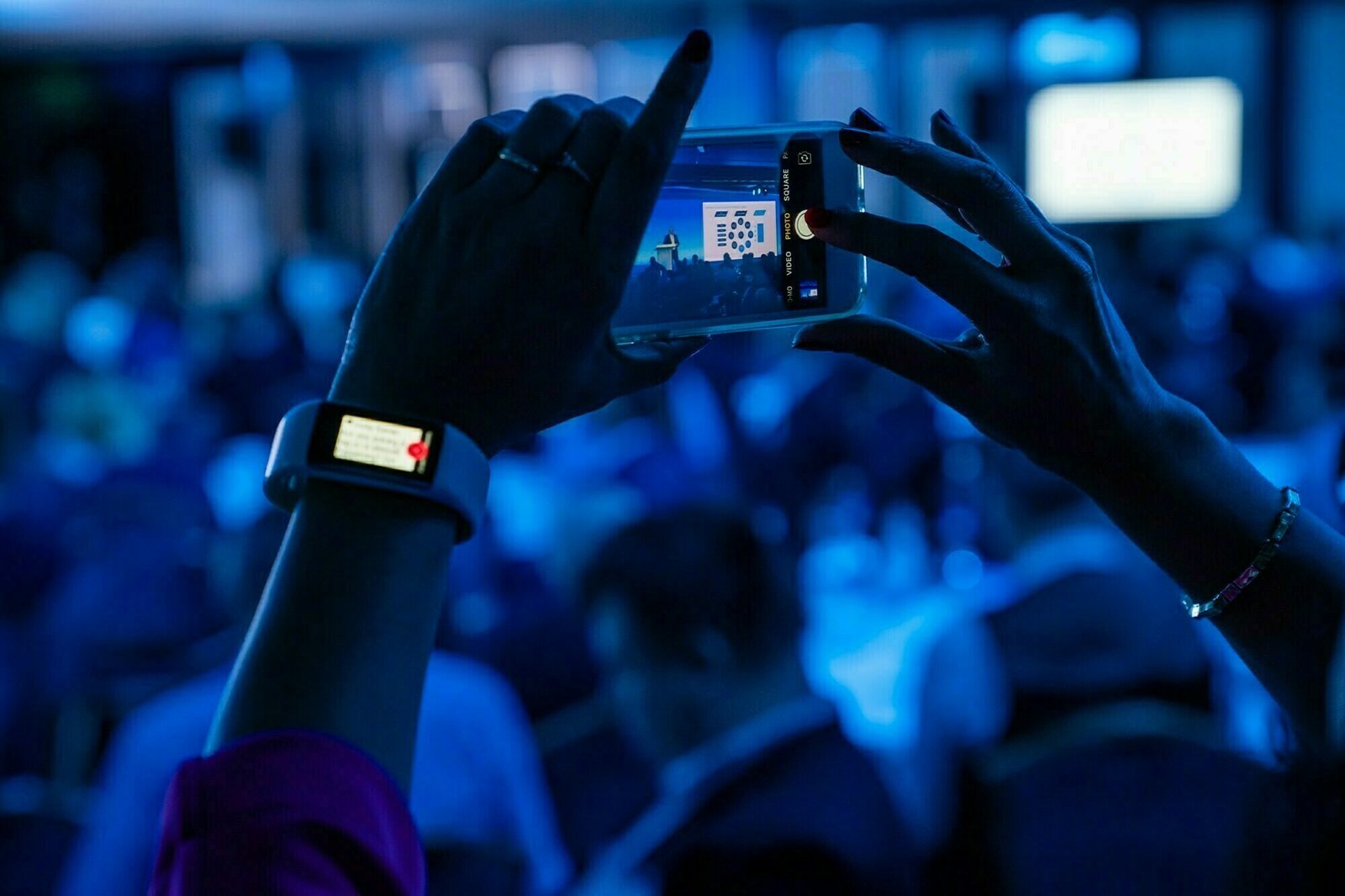 Close-up of a person taking a photo with a smartphone at a conference. The scene is bathed in blue light. The person is wearing a smartwatch, and the blurred background features an audience and a presentation screen. James Gifford-Mead Photography - Event Photographer London