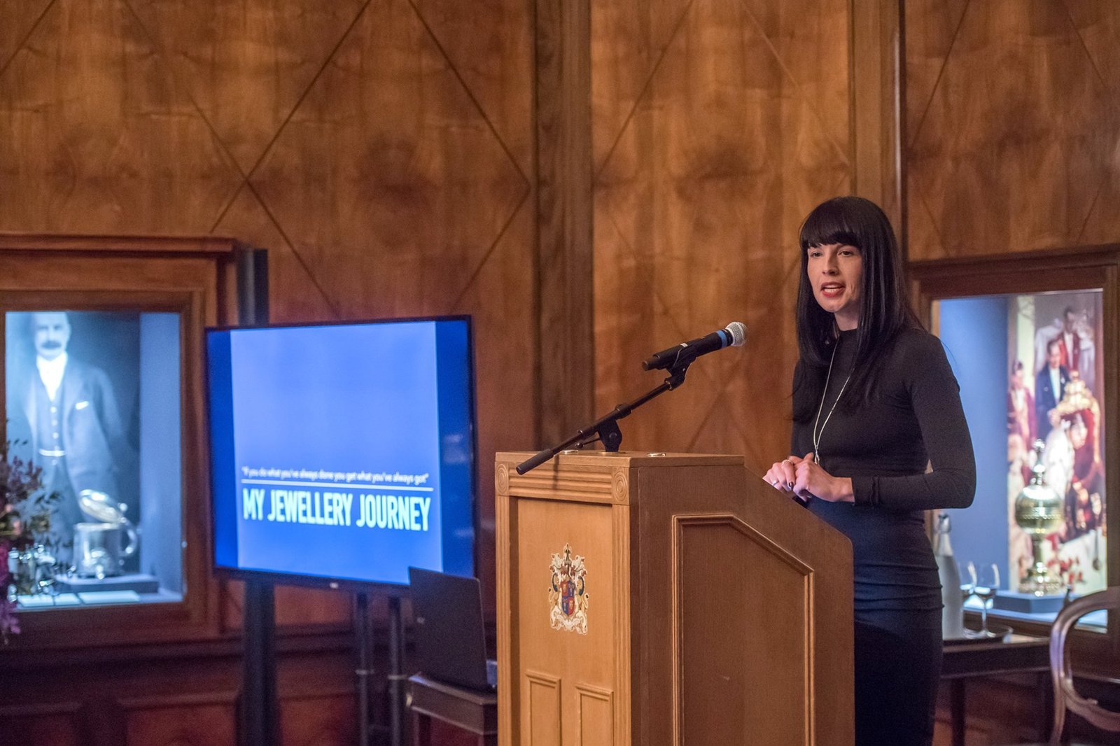 A woman stands at a podium speaking into a microphone in a wood-paneled room. Behind her, a screen displays "My Jewellery Journey" alongside vintage photographs and jewelry in display cases. James Gifford-Mead Photography - Event Photographer London