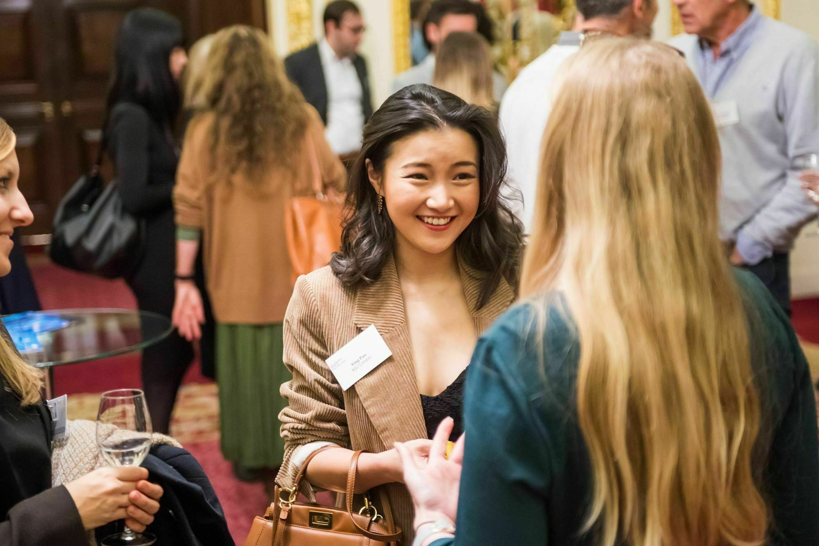 A smiling woman with a name tag is engaged in conversation with another woman at a networking event. She holds a glass of wine, and various people are gathered in the elegant, warmly lit room. James Gifford-Mead Photography - Event Photographer London