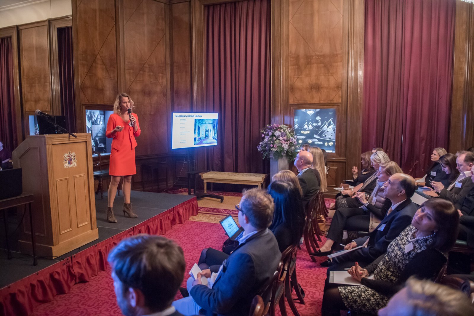 A woman in an orange dress is speaking at a podium in a richly decorated room with wooden paneling and red curtains. An audience is seated, listening attentively. A display screen shows a presentation slide next to the speaker