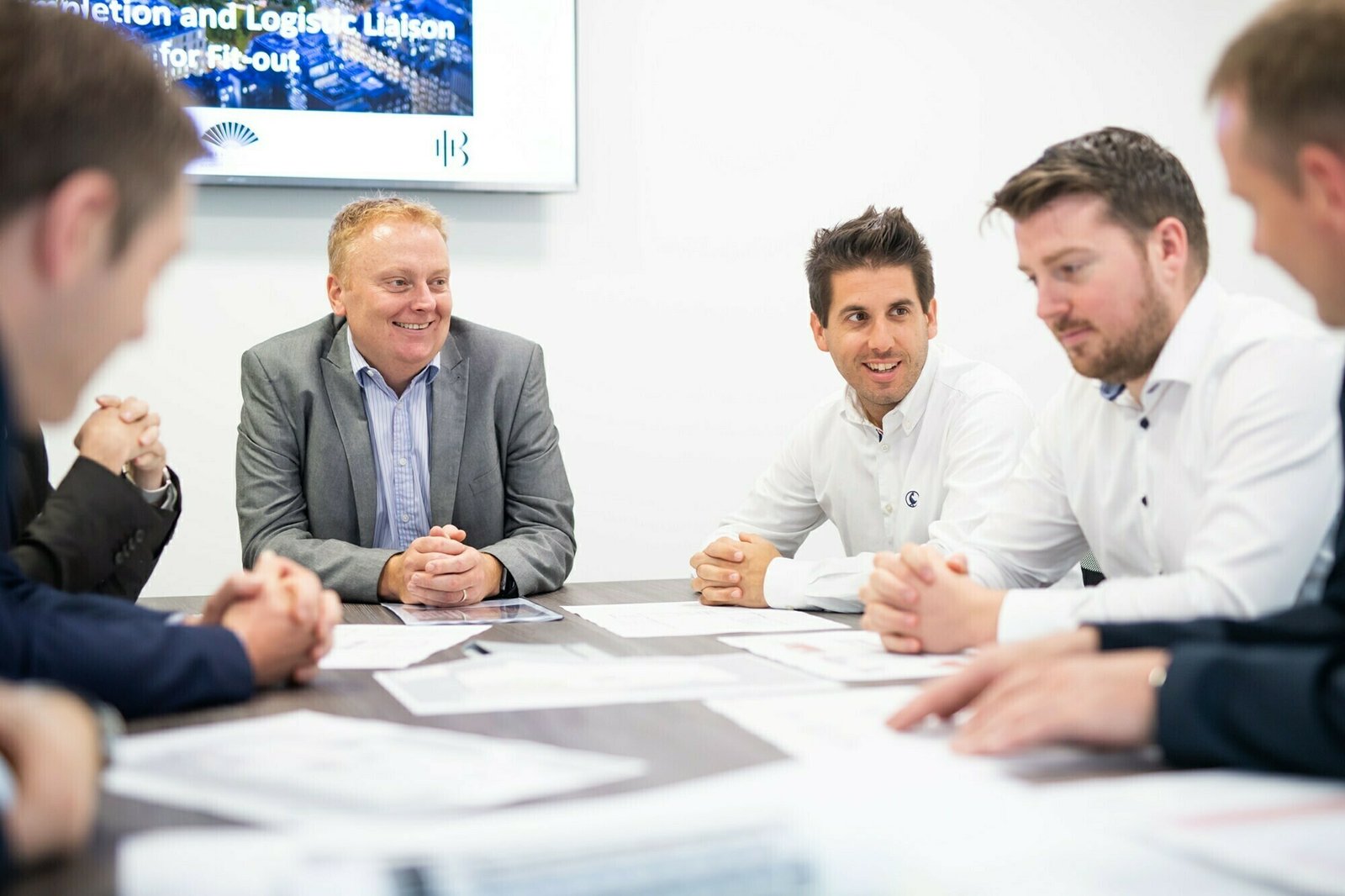 A group of six men are seated around a table in a meeting room, engaged in discussion. Some have papers in front of them. They appear attentive and are dressed in business attire. A screen in the background displays partially visible text. James Gifford-Mead Photography - Event Photographer London