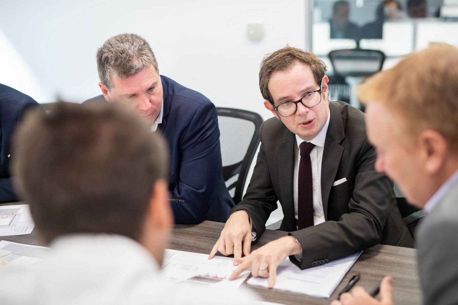 A group of four men in suits sit around a table engaged in discussion. One man gestures toward a document on the table. The setting appears to be a modern office with bright lighting. Papers and a laptop are visible on the table. James Gifford-Mead Photography - Event Photographer London