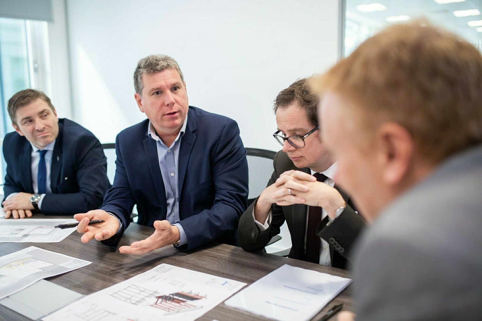 Four men in formal attire are seated around a table with documents. One man is speaking, gesturing with his hands, while the others listen attentively. The room appears to be an office setting with bright lighting. James Gifford-Mead Photography - Event Photographer London