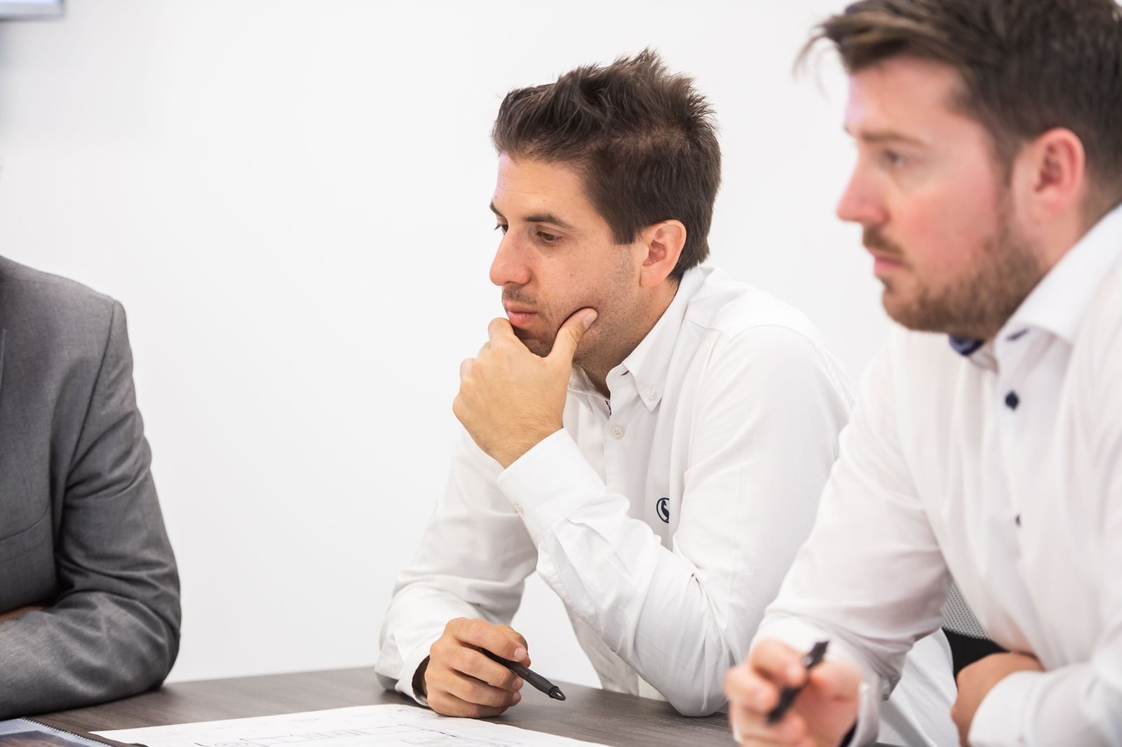 Three men seated at a table, engaged in a discussion. The man in focus, wearing a white shirt, rests his hand on his chin, appearing thoughtful. Papers are on the table, and two men hold pens. The setting is a bright conference room. James Gifford-Mead Photography - Event Photographer London