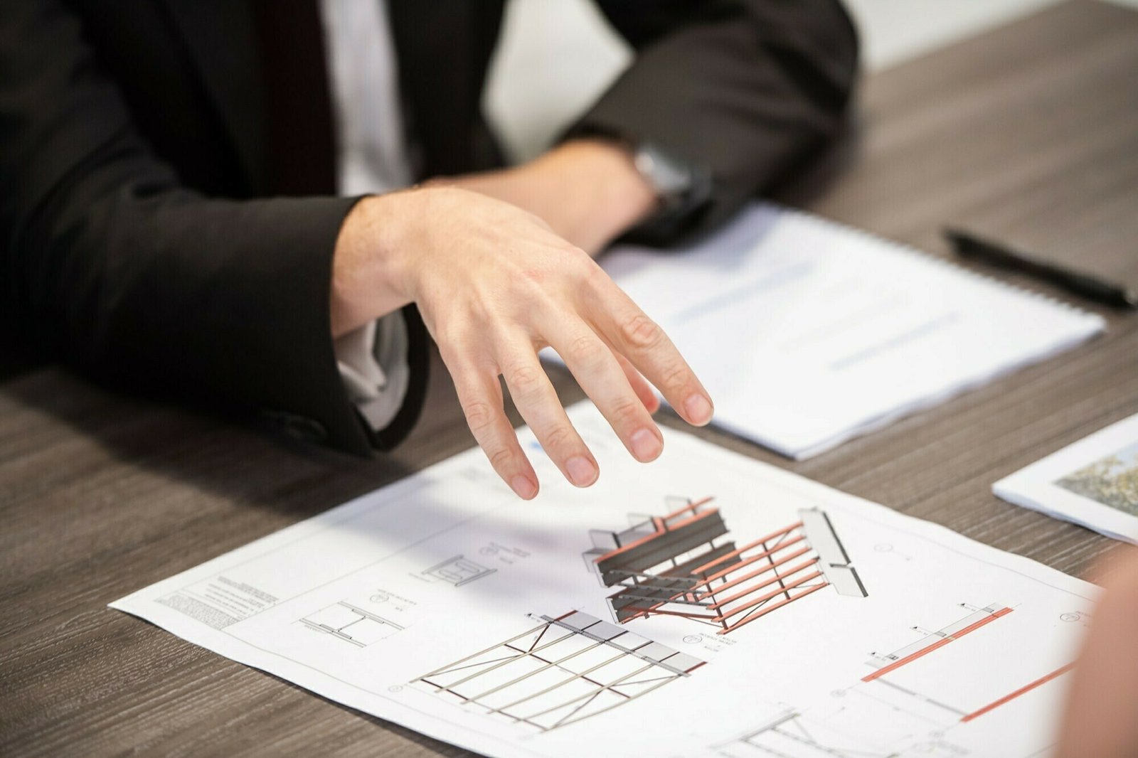 A person in a suit gestures over architectural plans spread on a wooden table. Various diagrams and designs are visible. A notebook and pen are placed nearby