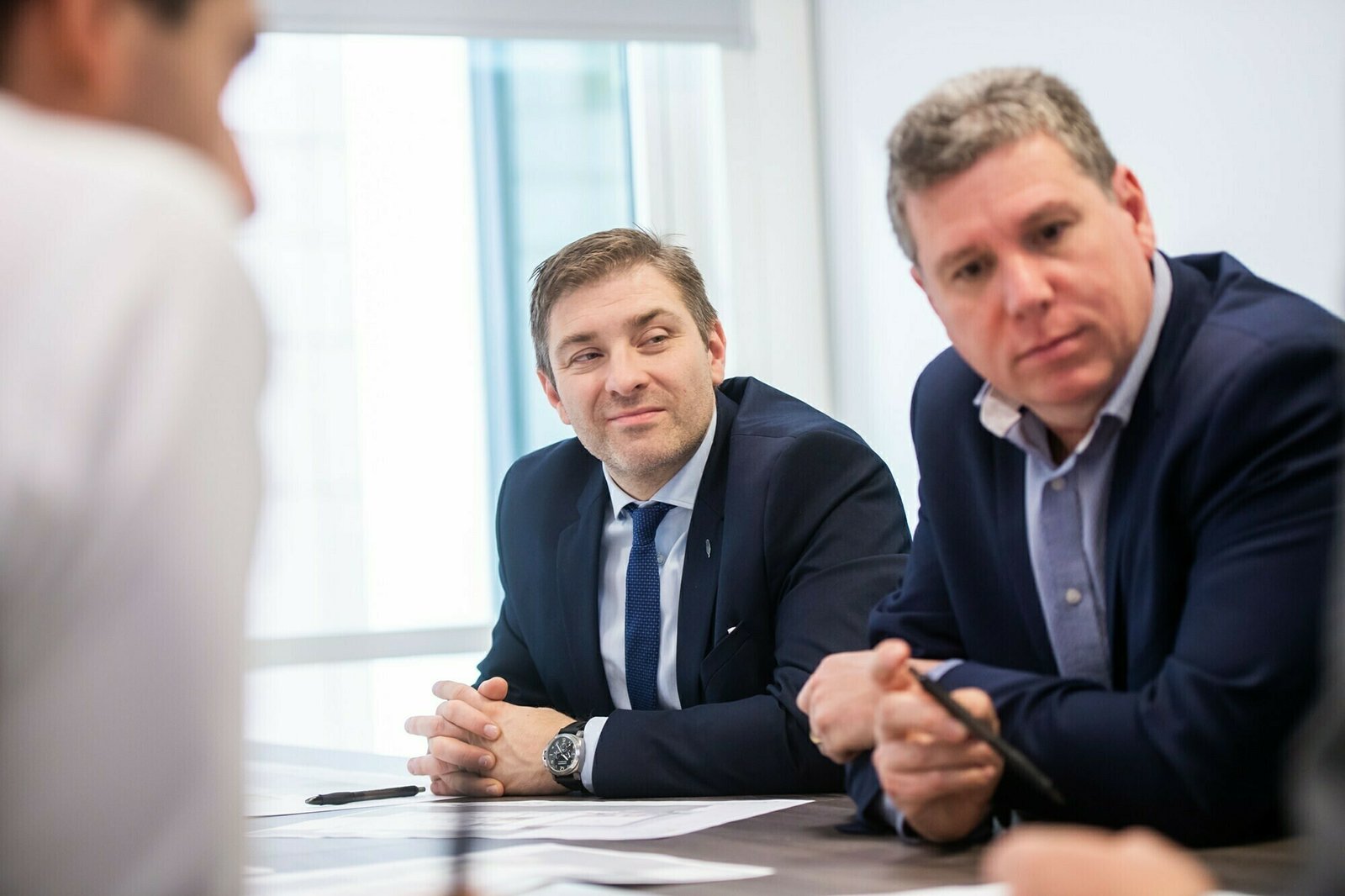 Two men in business attire are seated at a conference table engaged in discussion. One is looking attentively to the side, while the other, wearing a suit and tie, faces forward with a slight smile. A bright window is in the background. James Gifford-Mead Photography - Event Photographer London