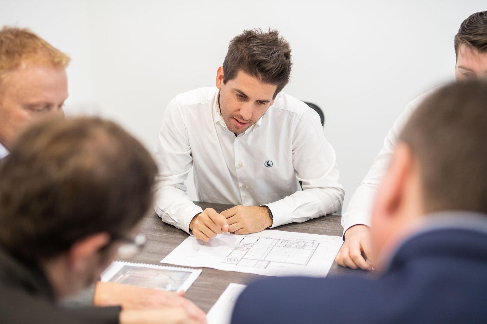 A group of people sitting around a table, discussing and examining architectural plans. A man in a white shirt is focused on the documents in front of him. The atmosphere appears to be professional and collaborative. James Gifford-Mead Photography - Event Photographer London