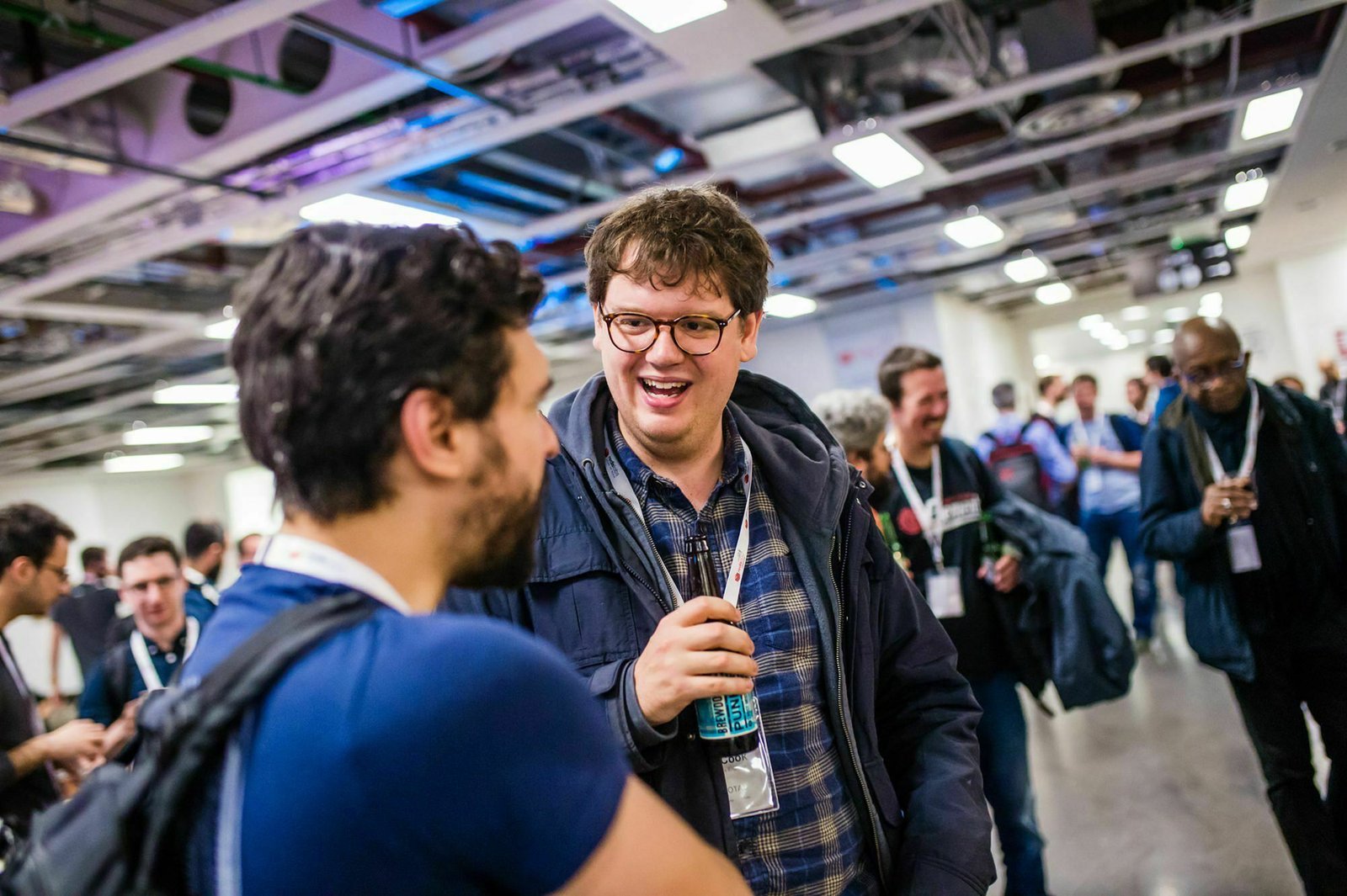 A group of people at an indoor event, with two men talking and smiling in the foreground. One holds a water bottle. Several others are visible in the background, mingling and conversing. The setting is informal, with visible ceiling pipes. James Gifford-Mead Photography - Event Photographer London