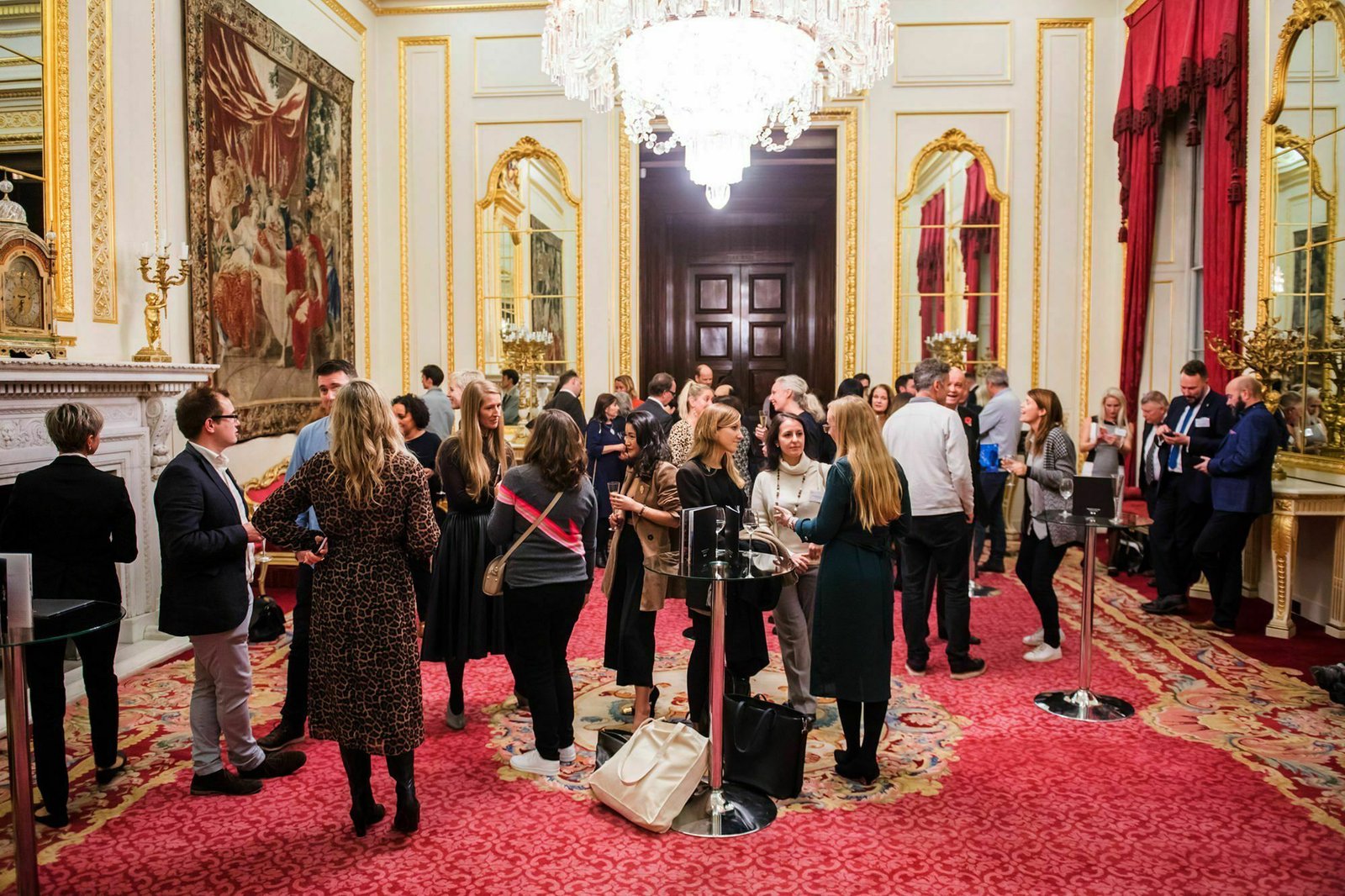 A group of people mingling in an ornate room with gold-framed mirrors, a large chandelier, and red carpet. The walls are adorned with tapestries and a fireplace. Several small tables are scattered around the room. James Gifford-Mead Photography - Event Photographer London