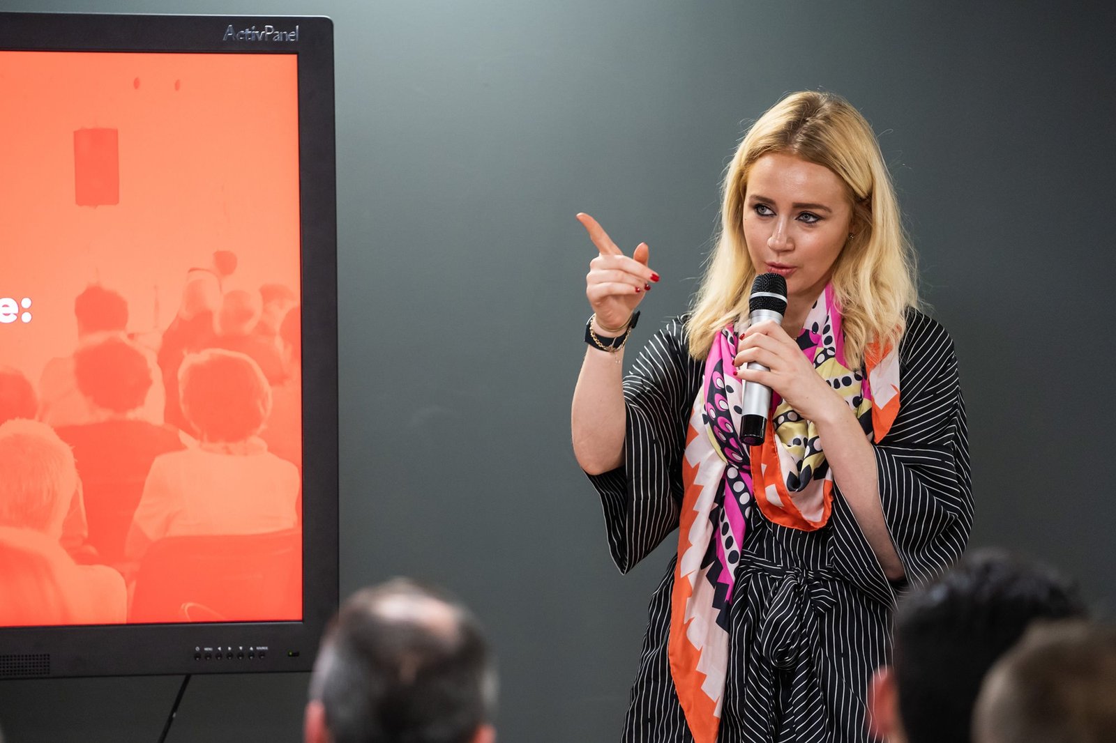 A woman with blonde hair speaks into a microphone while gesturing with her hand. She stands in front of an audience and a large screen with an orange background. She wears a colorful scarf and a striped outfit. James Gifford-Mead Photography - Event Photographer London