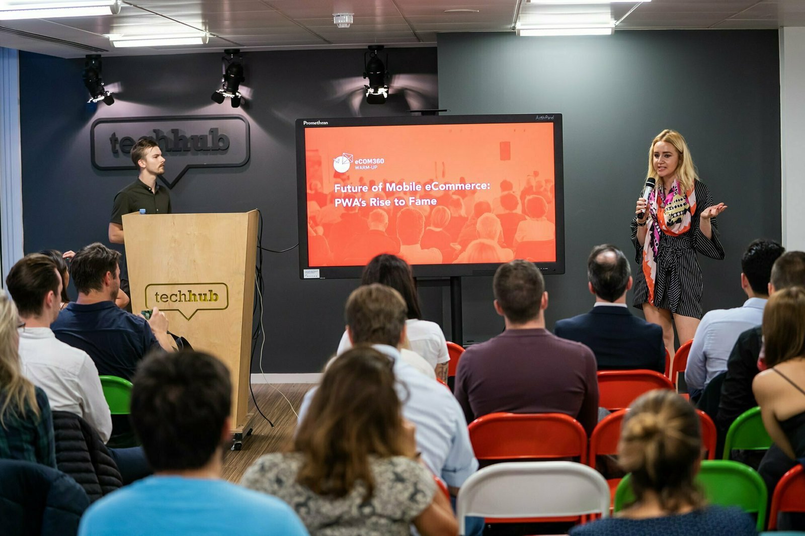 A woman speaks to an audience during a presentation on "Future of Mobile eCommerce: PWA's Rise to Fame" at a tech event. She stands next to a large screen displaying the presentation title. Attendees are seated, listening attentively. James Gifford-Mead Photography - Event Photographer London
