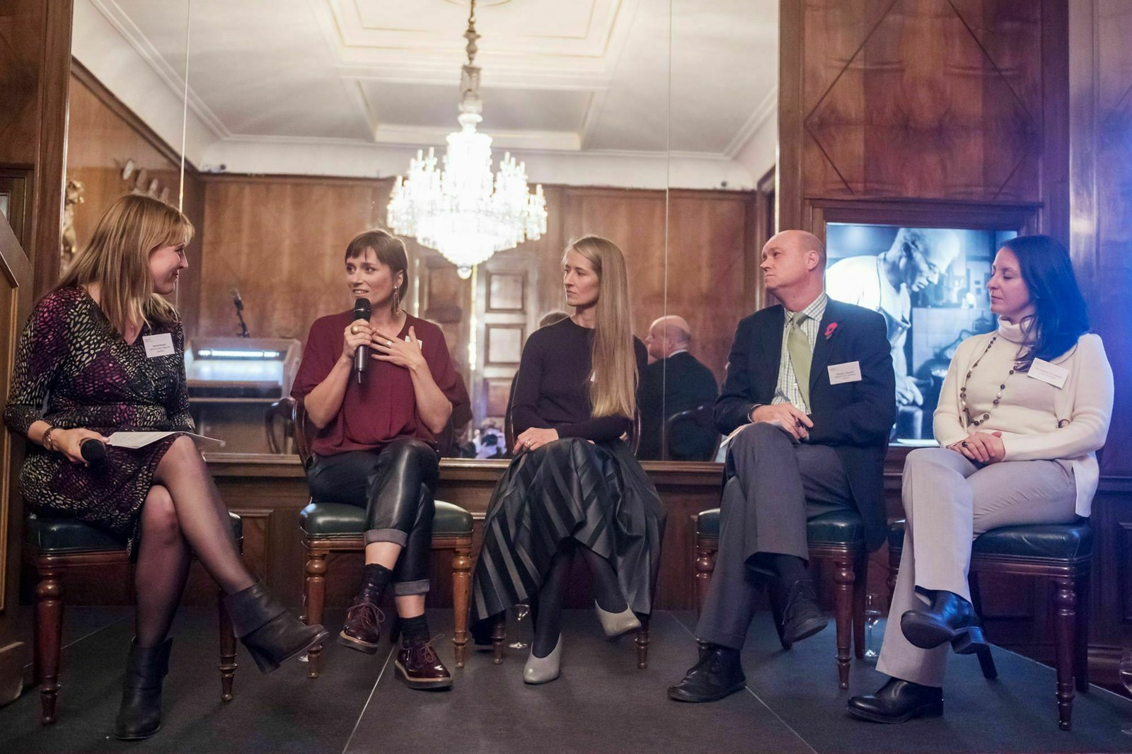 A panel of five individuals sitting and having a discussion in a warmly lit room with wooden walls and a chandelier. Three women are on the left, with one using a microphone, and two men are on the right, all dressed in business casual. James Gifford-Mead Photography - Event Photographer London