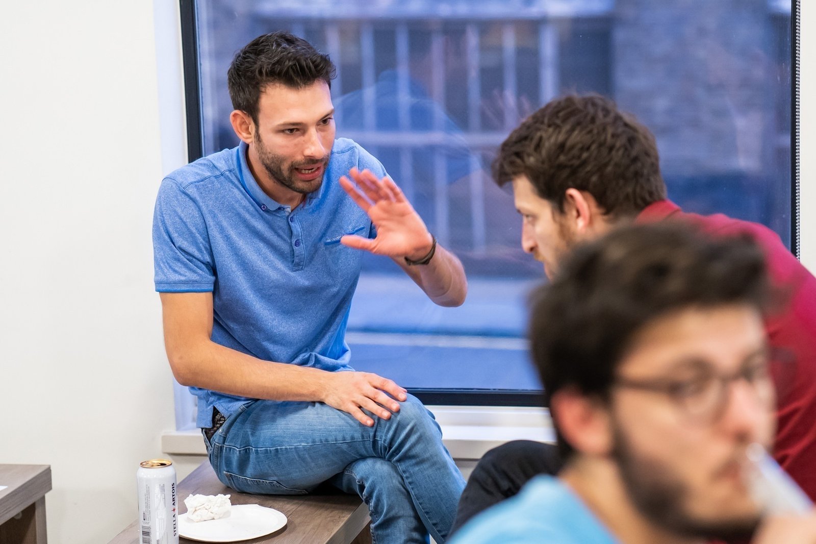 A man in a blue polo shirt is sitting on a windowsill, gesturing with his hand while talking to a man in a red shirt. Another person is blurred in the foreground. A plate and a can are on the windowsill. James Gifford-Mead Photography - Event Photographer London