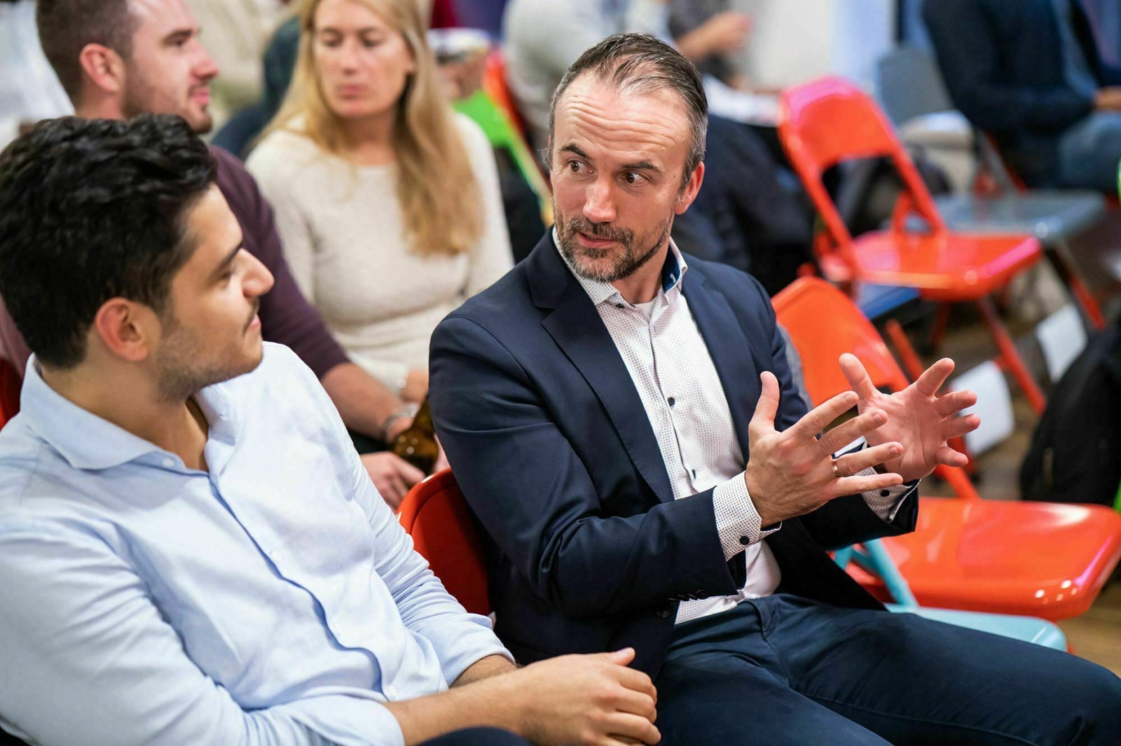Two men in a discussion sit on red chairs. The man on the right gestures with both hands while speaking to the man on the left, who listens attentively. Other people are seated in the background. James Gifford-Mead Photography - Event Photographer London